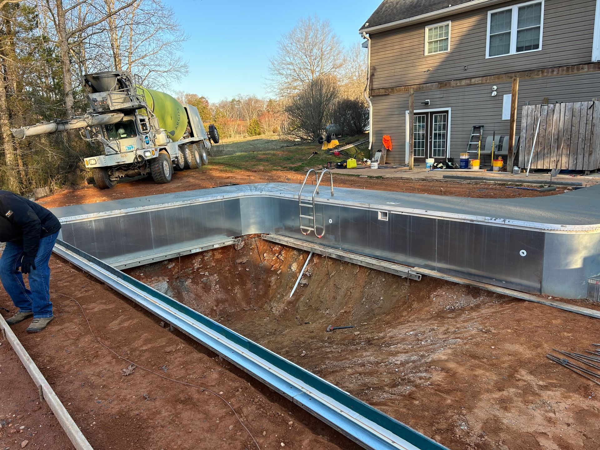 Construction site with a partially built pool. A cement truck pours cement. A worker is near the pool's edge.