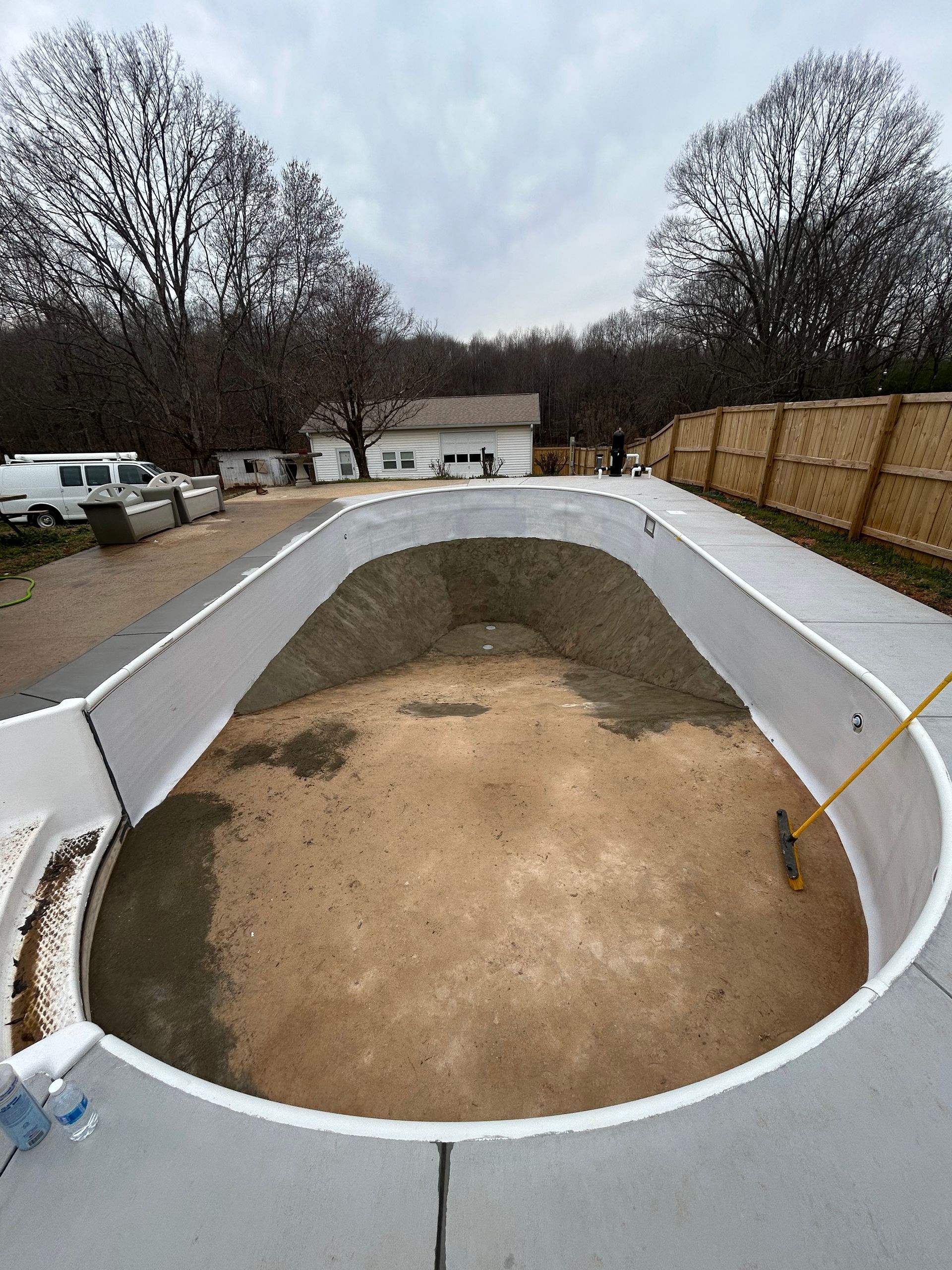 Empty oval swimming pool under construction, concrete surrounding with a wooden fence in the background.
