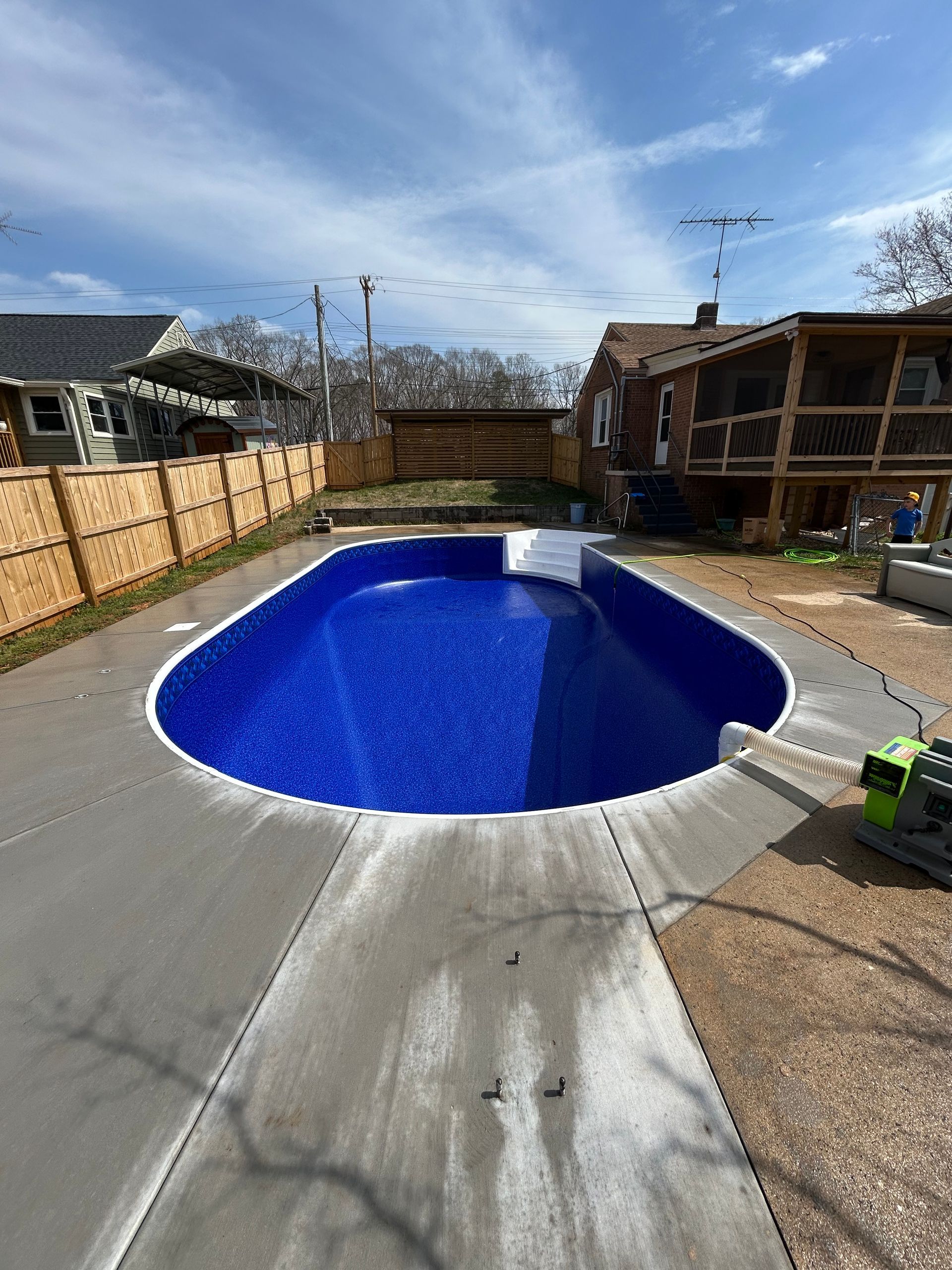 Oval-shaped swimming pool with blue interior and concrete surround, near fenced yard and houses.