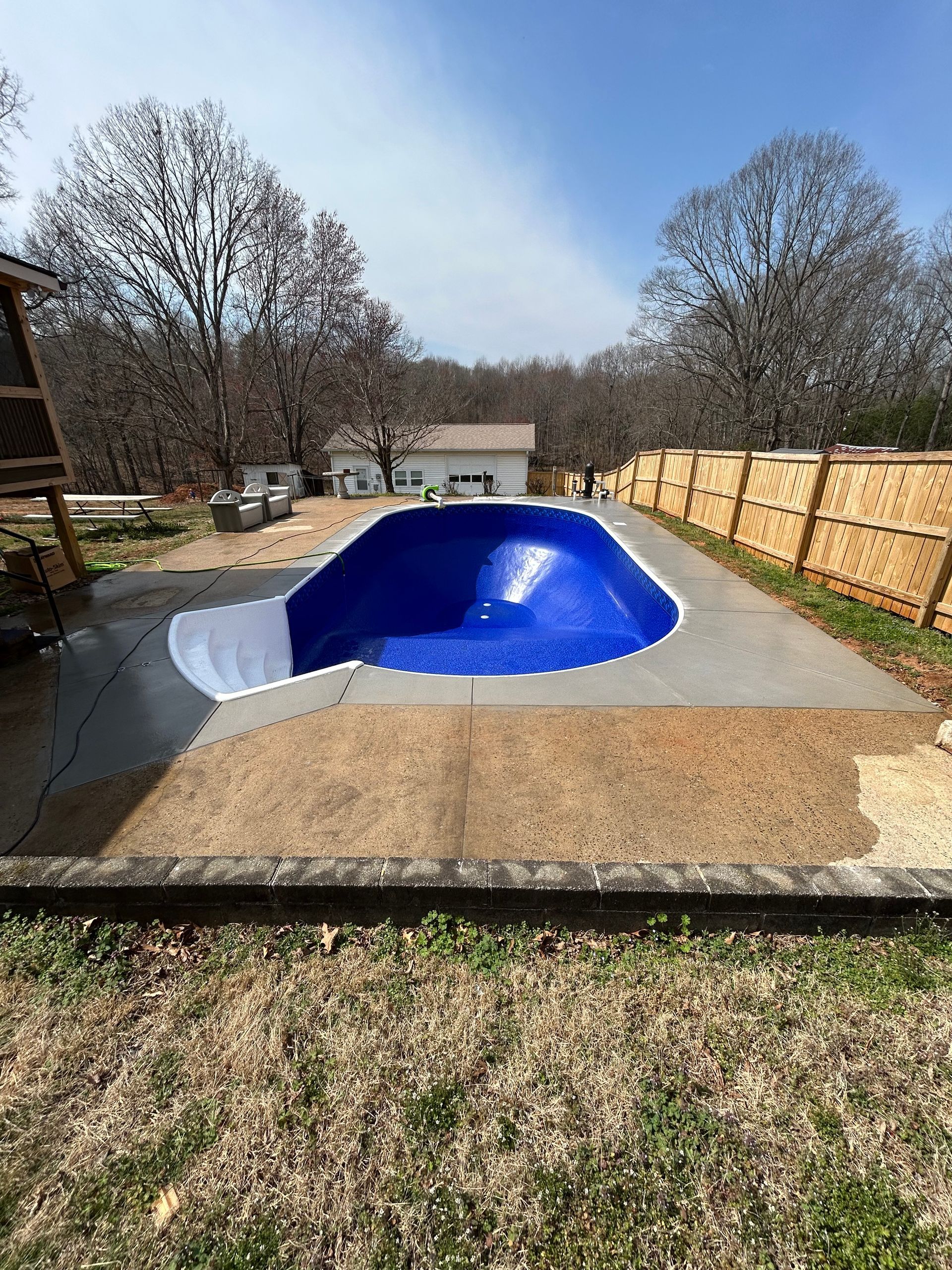 A partially filled blue swimming pool with gray concrete decking.  A wooden fence and trees surround the pool.