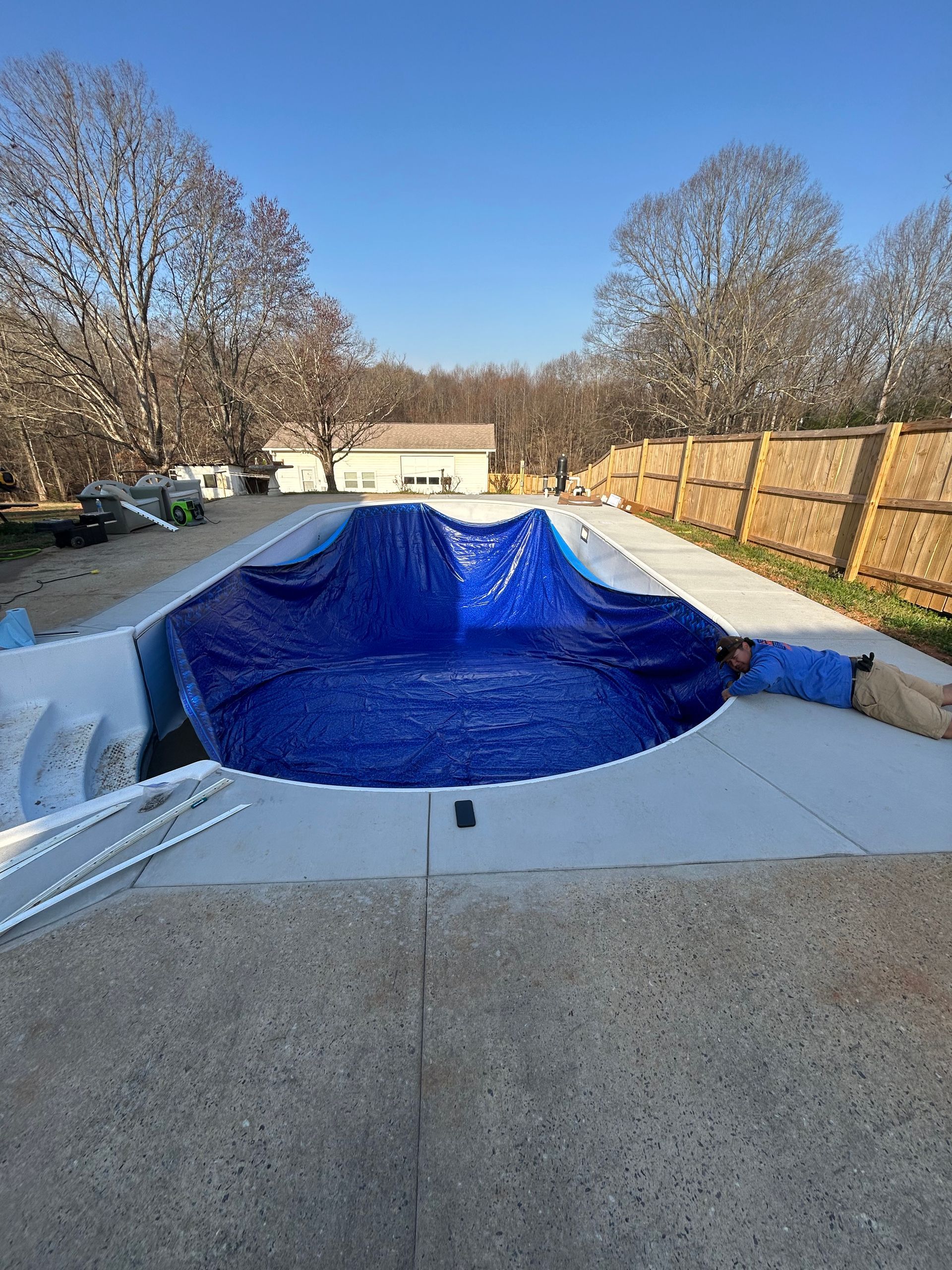 Blue tarp covers a concrete pool. Man lays beside it on the right. Trees, fence, and clear sky in background.