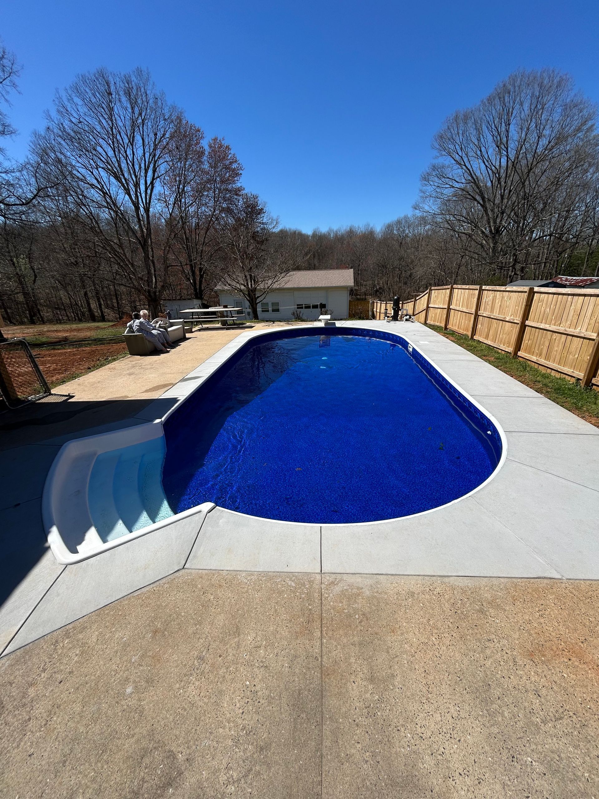 Blue-tiled pool with concrete surround and steps, under a clear blue sky. Trees and a building in the background.