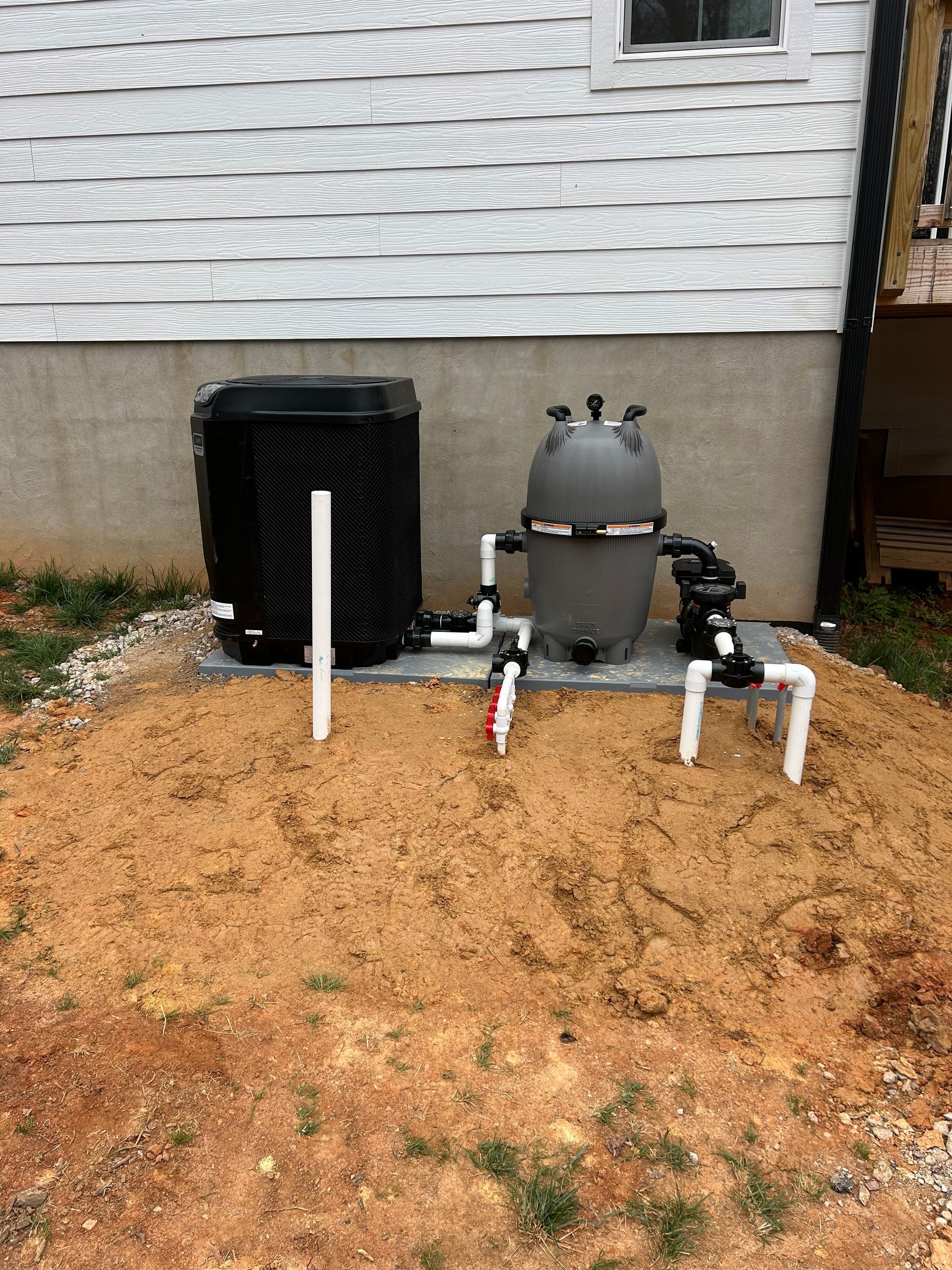 Pool equipment: filter, heater, and plumbing installed on a sand bed, next to a white house.
