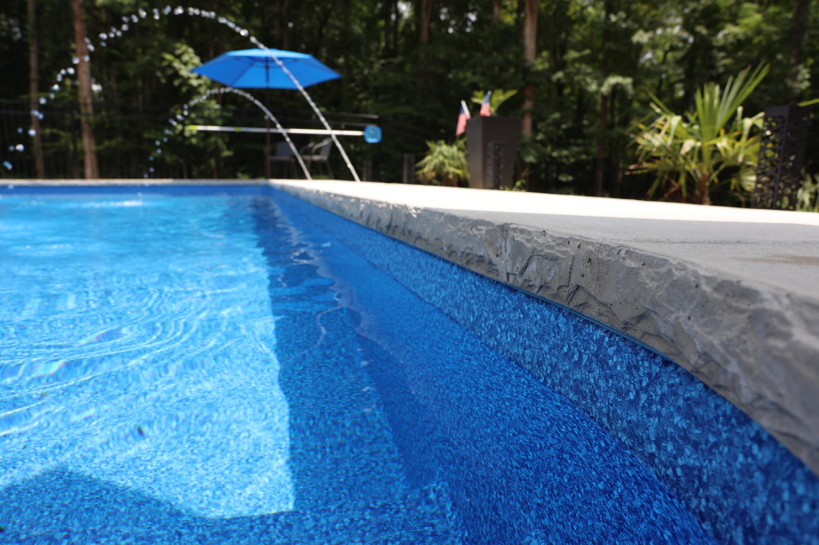 Close-up of a swimming pool with a blue interior and a grey stone edge. Water is splashing and a blue umbrella is visible.