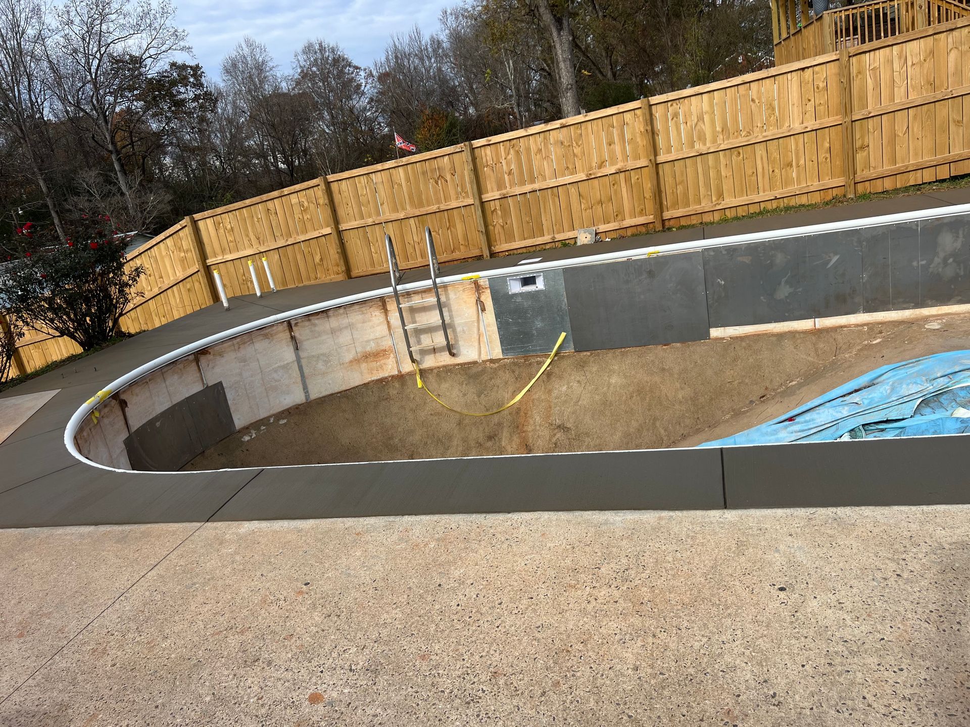Empty, in-ground swimming pool with concrete deck and wooden fence; ladder and plumbing visible.
