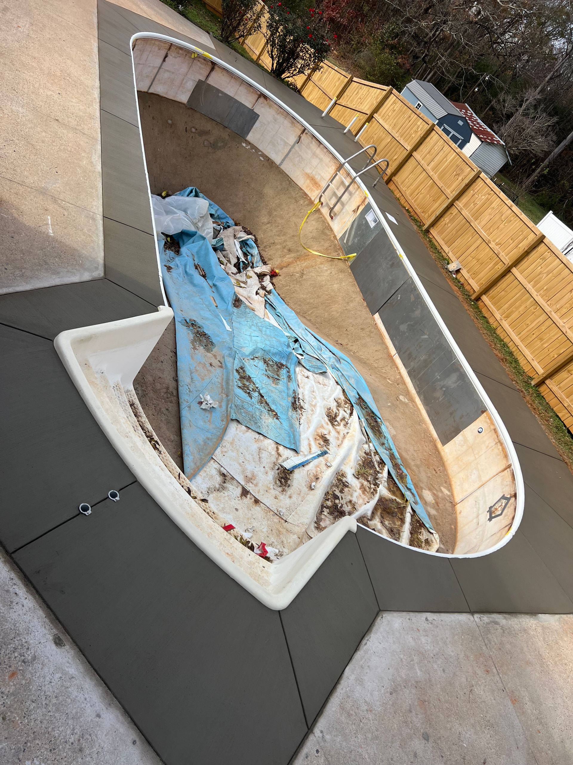 Empty pool with gray concrete surround, debris in the basin, and a wood fence.