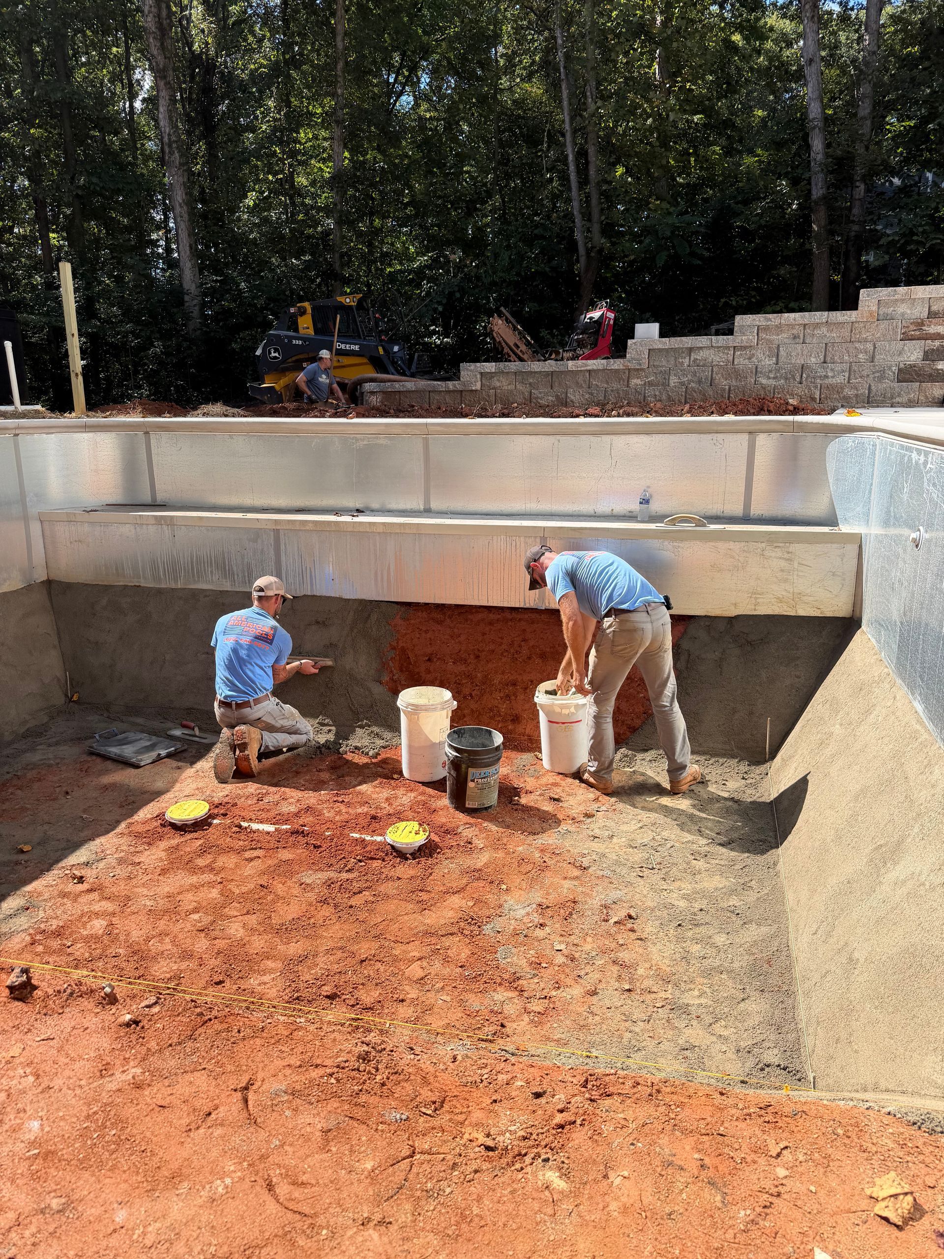 Two workers in a pool construction site applying material; one kneels, the other stands.