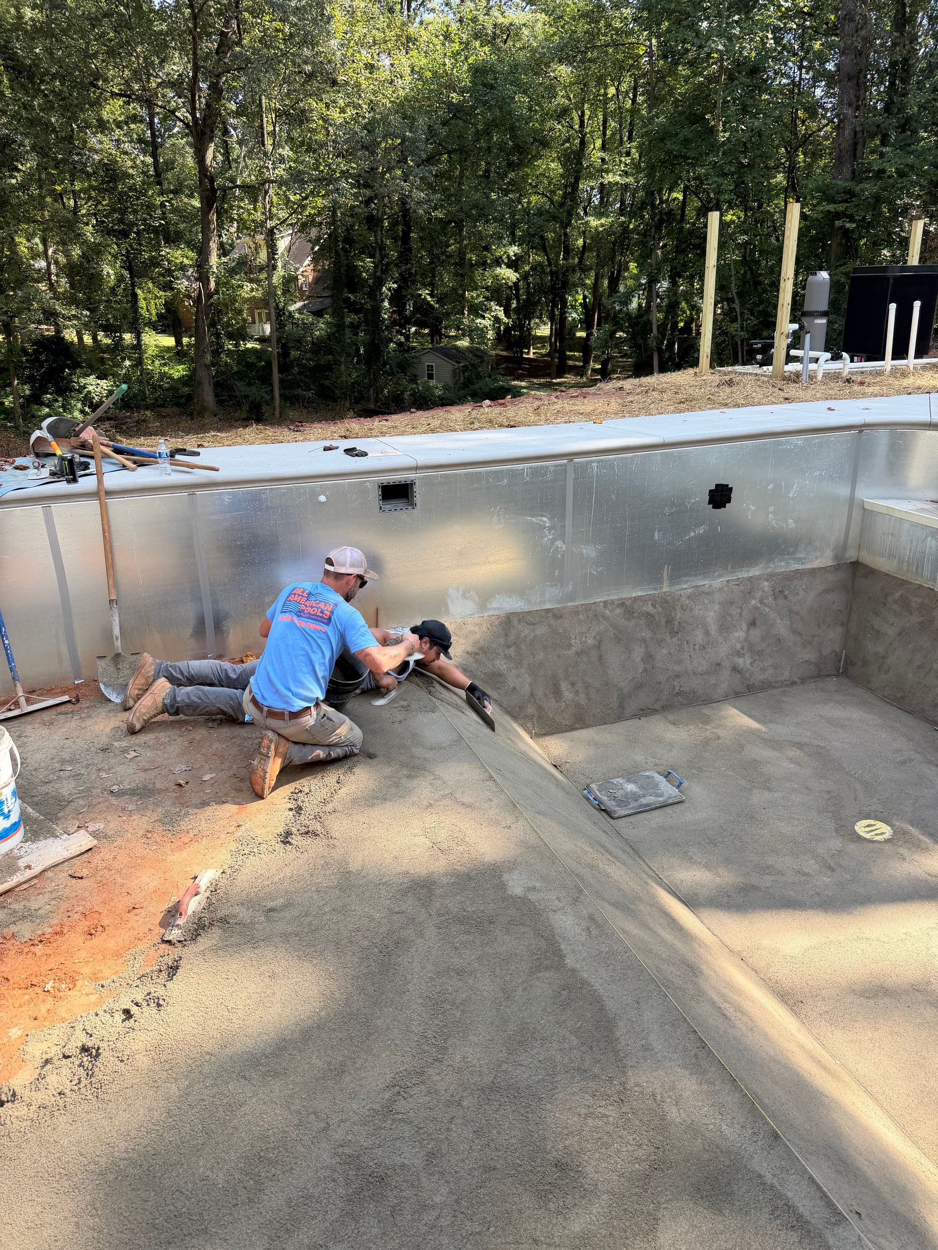 Two workers smoothing concrete inside a partially built swimming pool. Sunny outdoors with trees in the background.