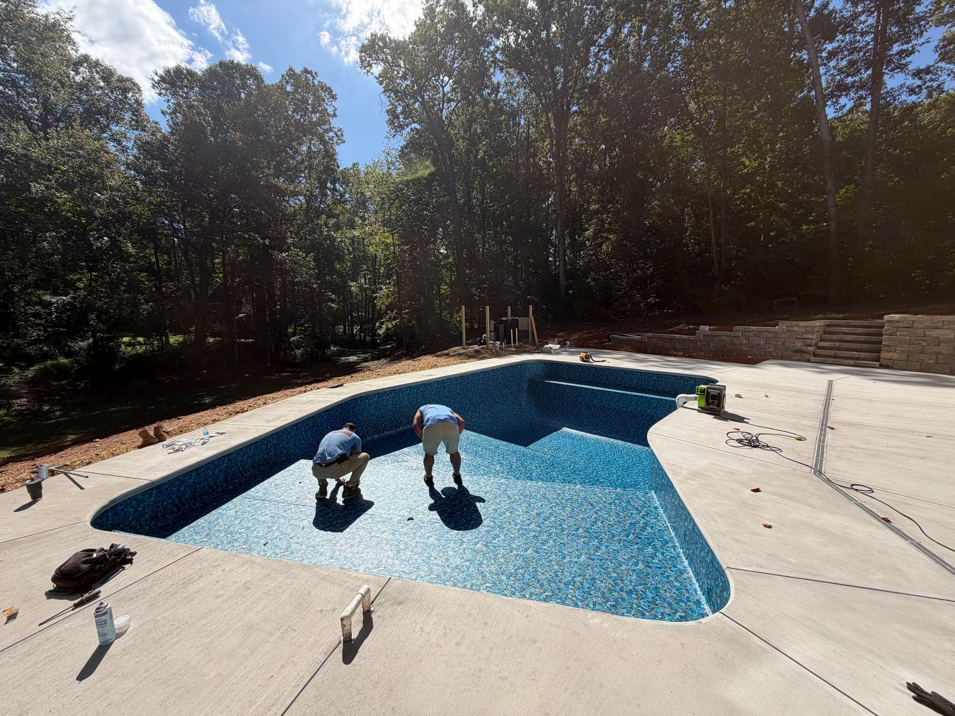 Workers installing blue tile in a newly built swimming pool. Concrete patio, trees in background. Sunny day.