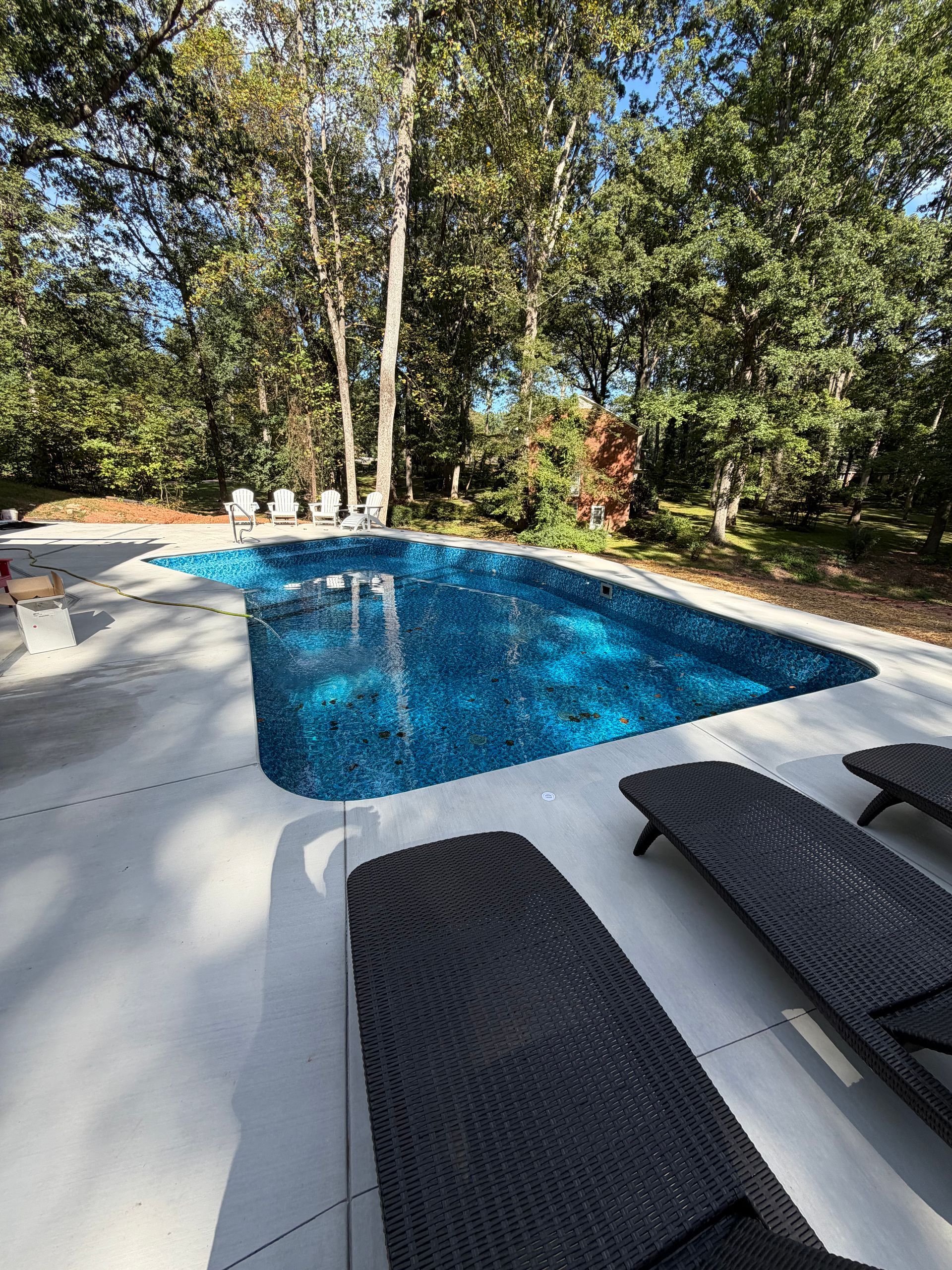 Swimming pool with blue water surrounded by a white patio and lounge chairs. Trees in the background.