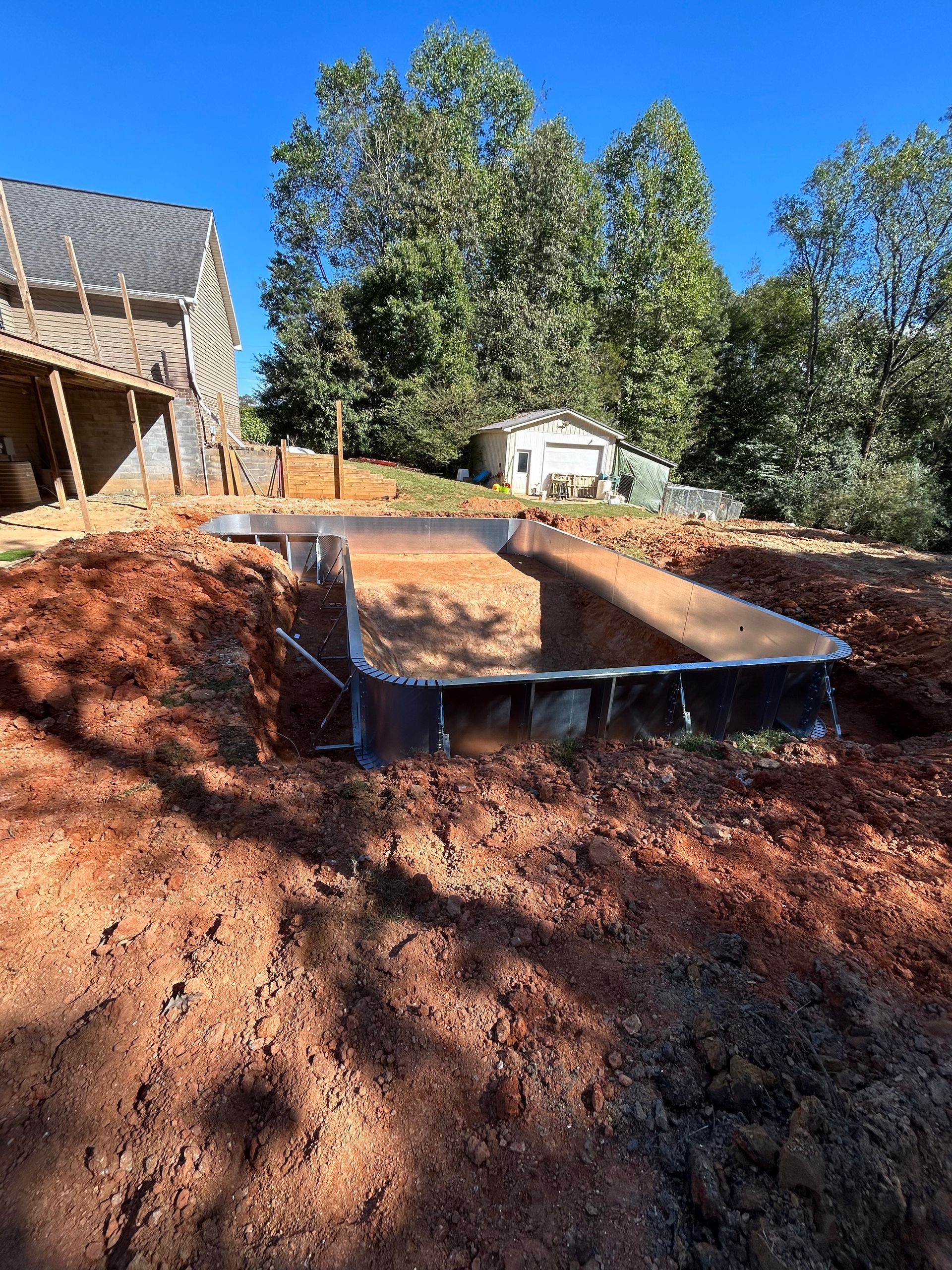 Rectangular pool under construction in a dirt yard, house in background, clear blue sky.