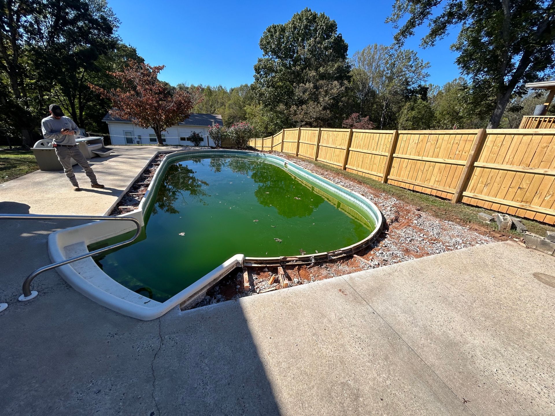 Pool filled with green water needing repair, person standing nearby, wooden fence in the background, sunny day.