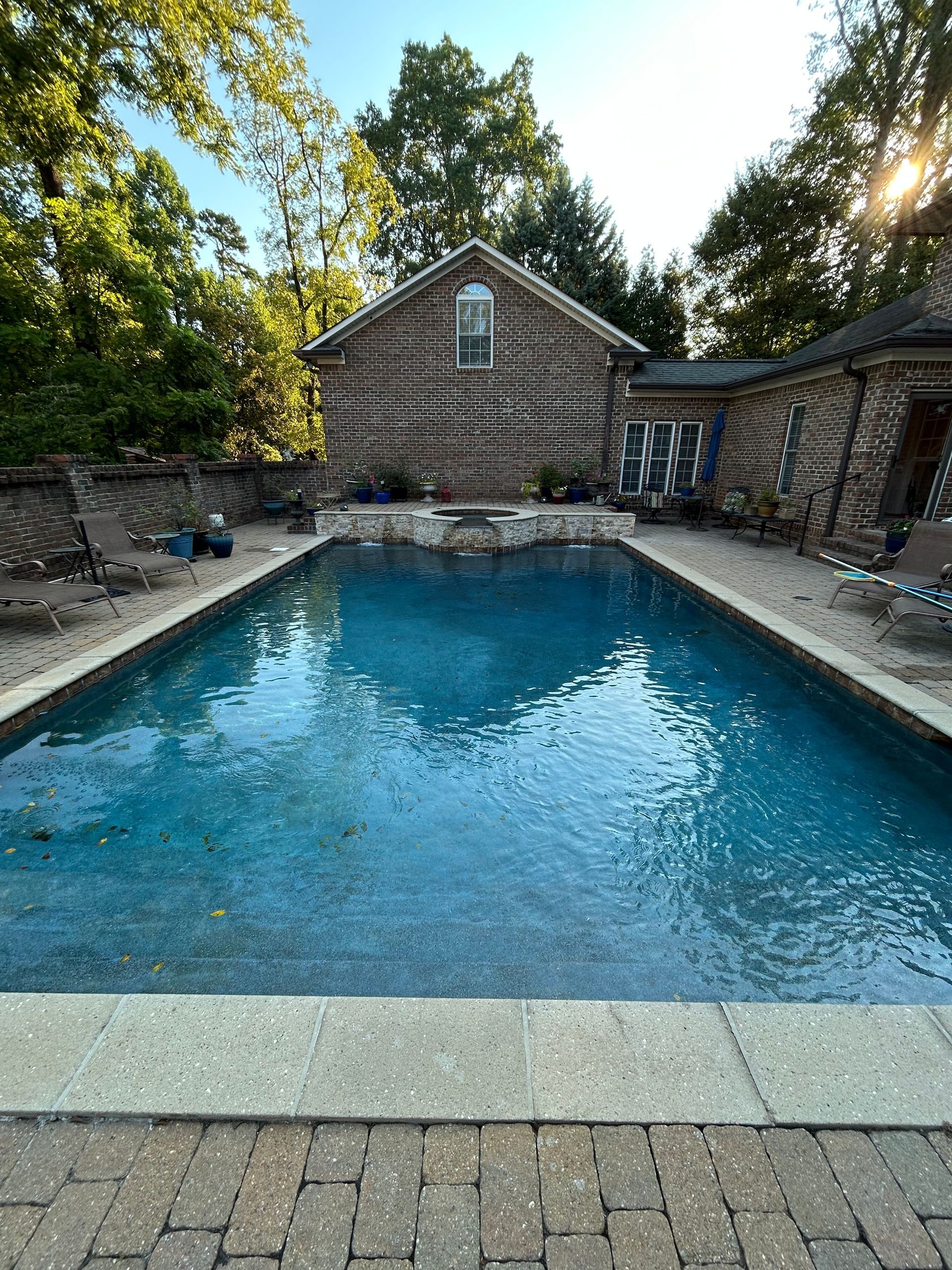 A rectangular swimming pool with clear blue water, surrounded by brick and stone, with a brick building in the background.