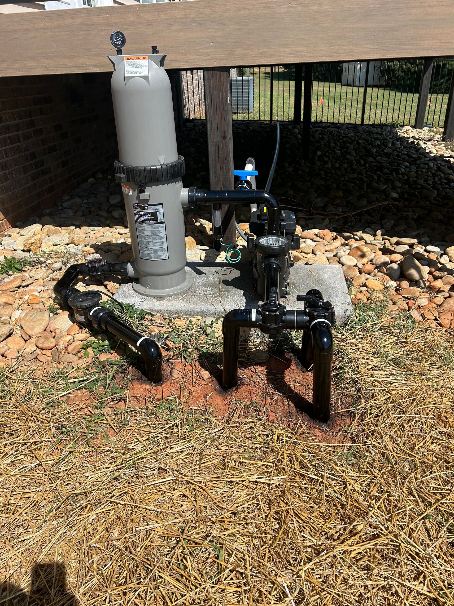 Pool filter system on a concrete base, with black pipes, a gray filter, and a pressure gauge.