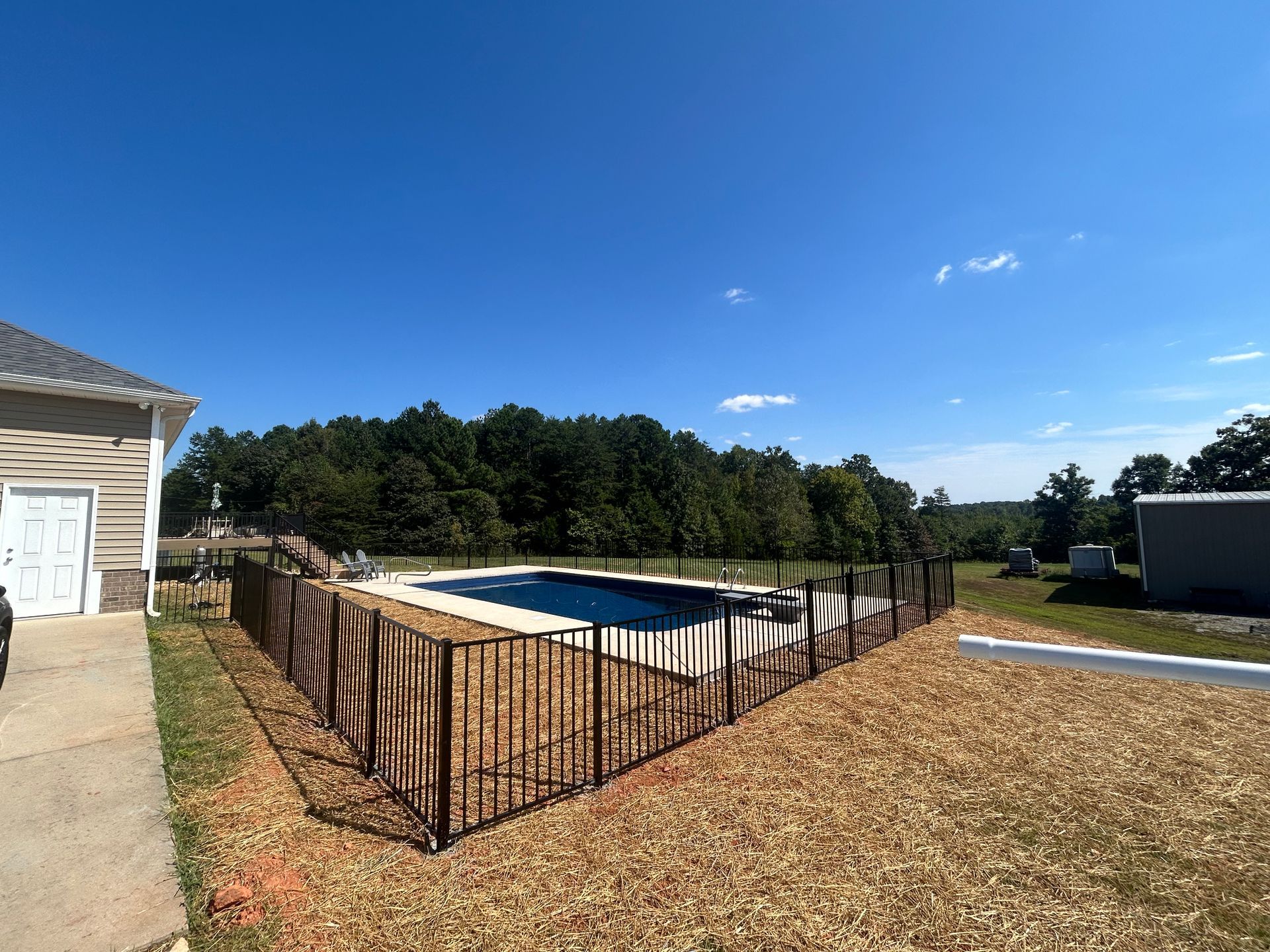 A fenced-in rectangular pool on a sunny day. The fence is dark with a pattern. Buildings and trees are in the background.