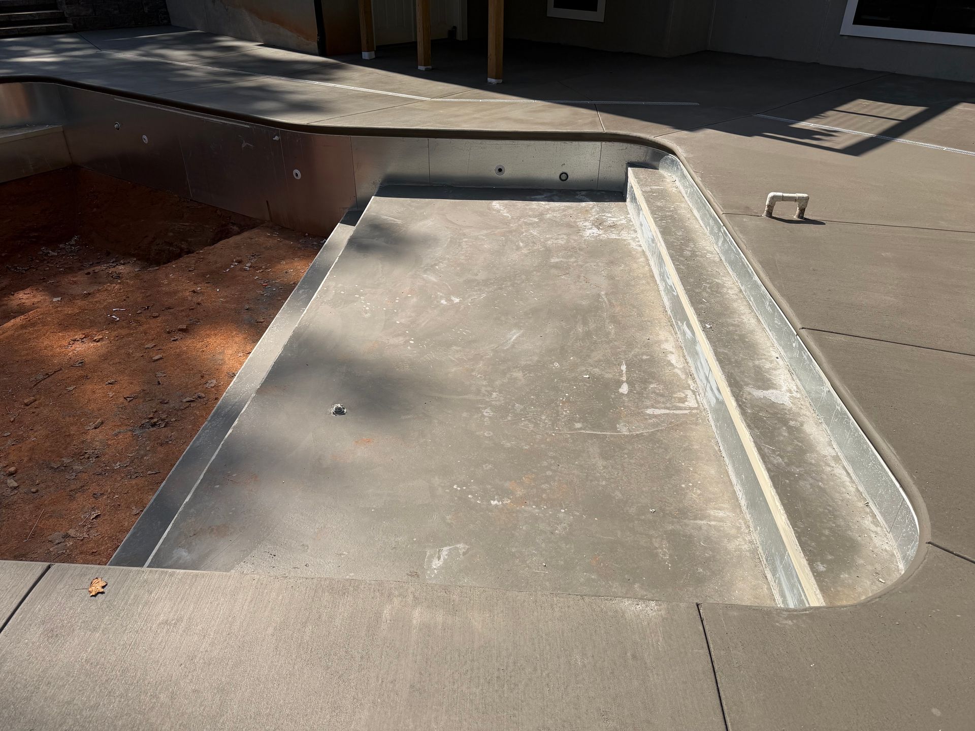 Empty, concrete swimming pool with steps. Brown dirt visible in the deep end, gray patio surrounding.