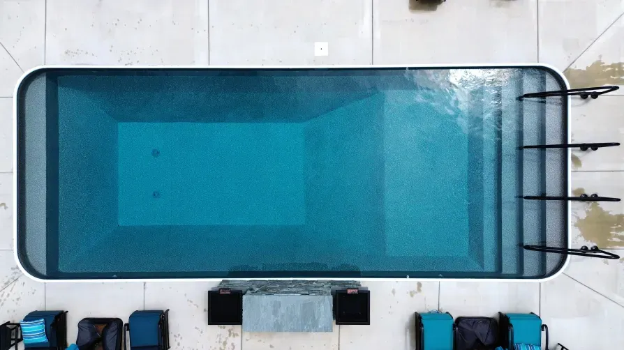 Overhead view of a rectangular turquoise pool with stairs, surrounded by white stone and dark blue equipment.
