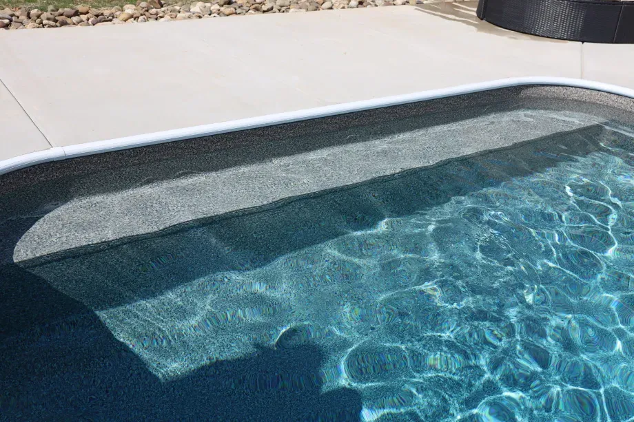 Pool with built-in steps, reflecting sunlight. Concrete deck surrounds the pool.