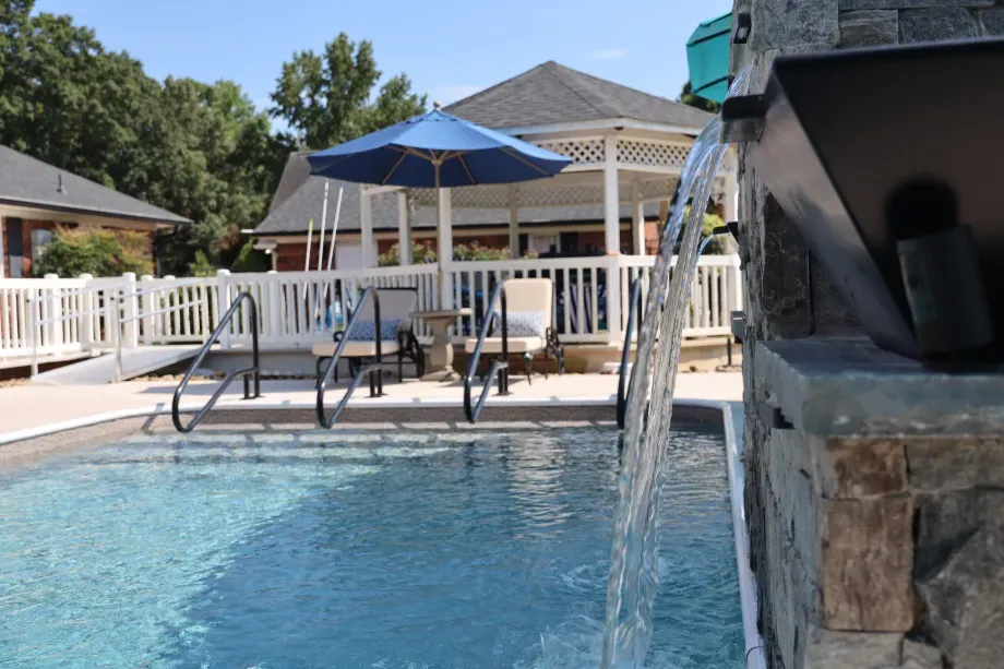 Pool with a waterfall, gazebo, umbrella, and chairs. Blue water and sky.