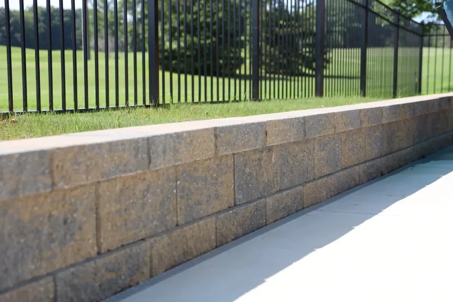 Low retaining wall of brown blocks next to a grassy area, a fence, and a concrete surface.
