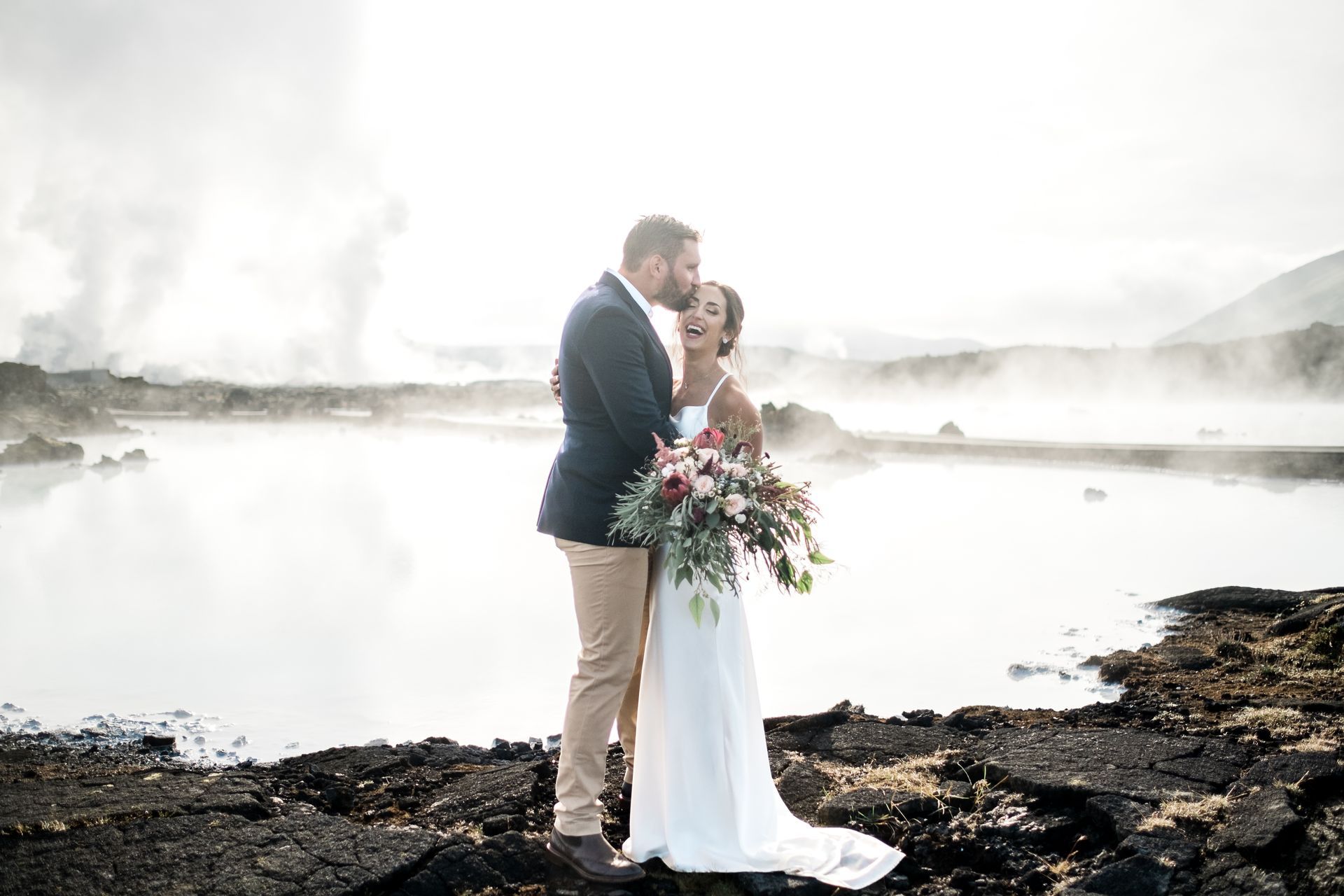A bride and groom are standing next to each other on the shore of a lake.