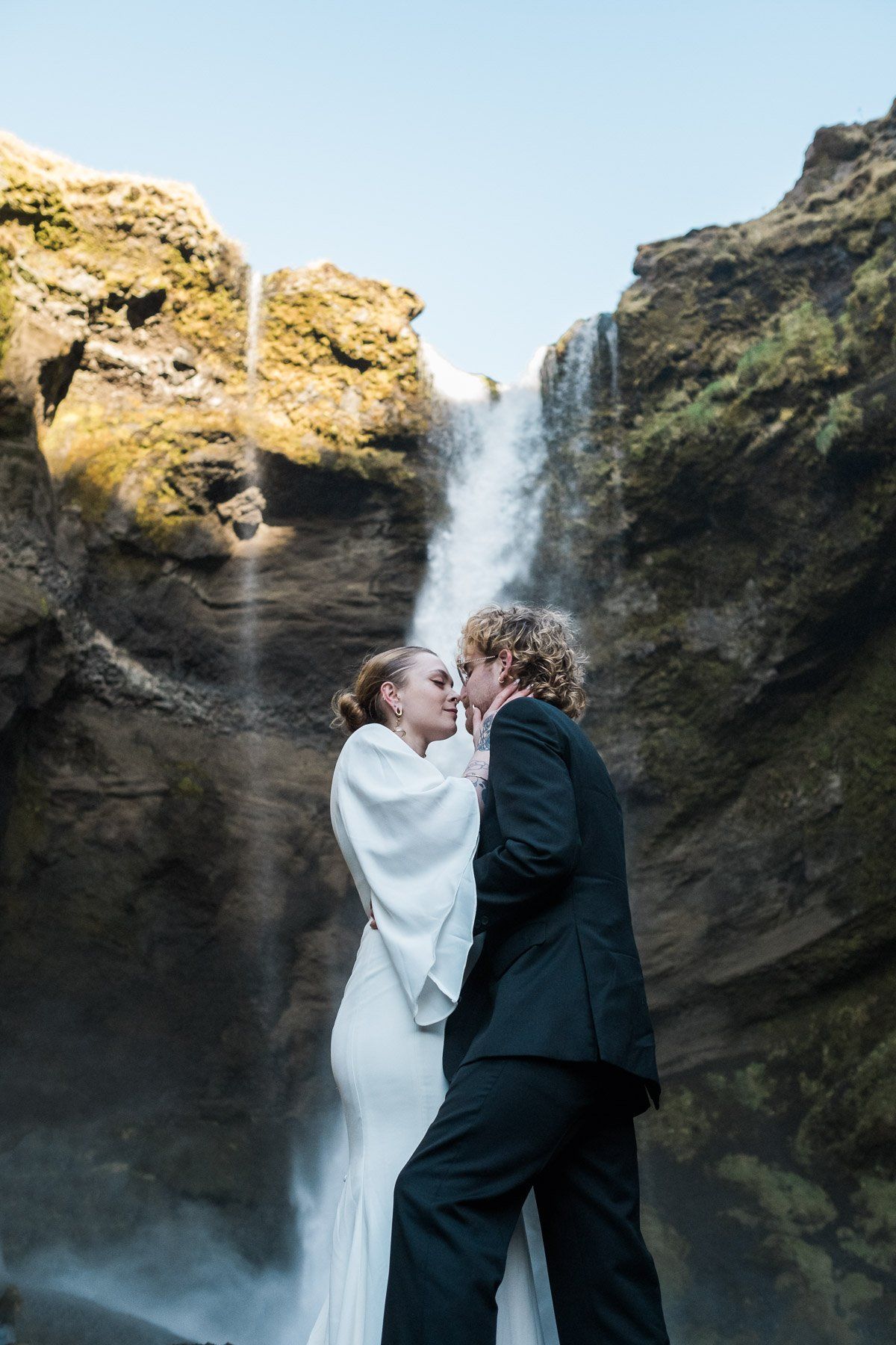 A couple shares a romantic moment by a waterfall during their Iceland elopement, framed by cliffs and a clear blue sky.