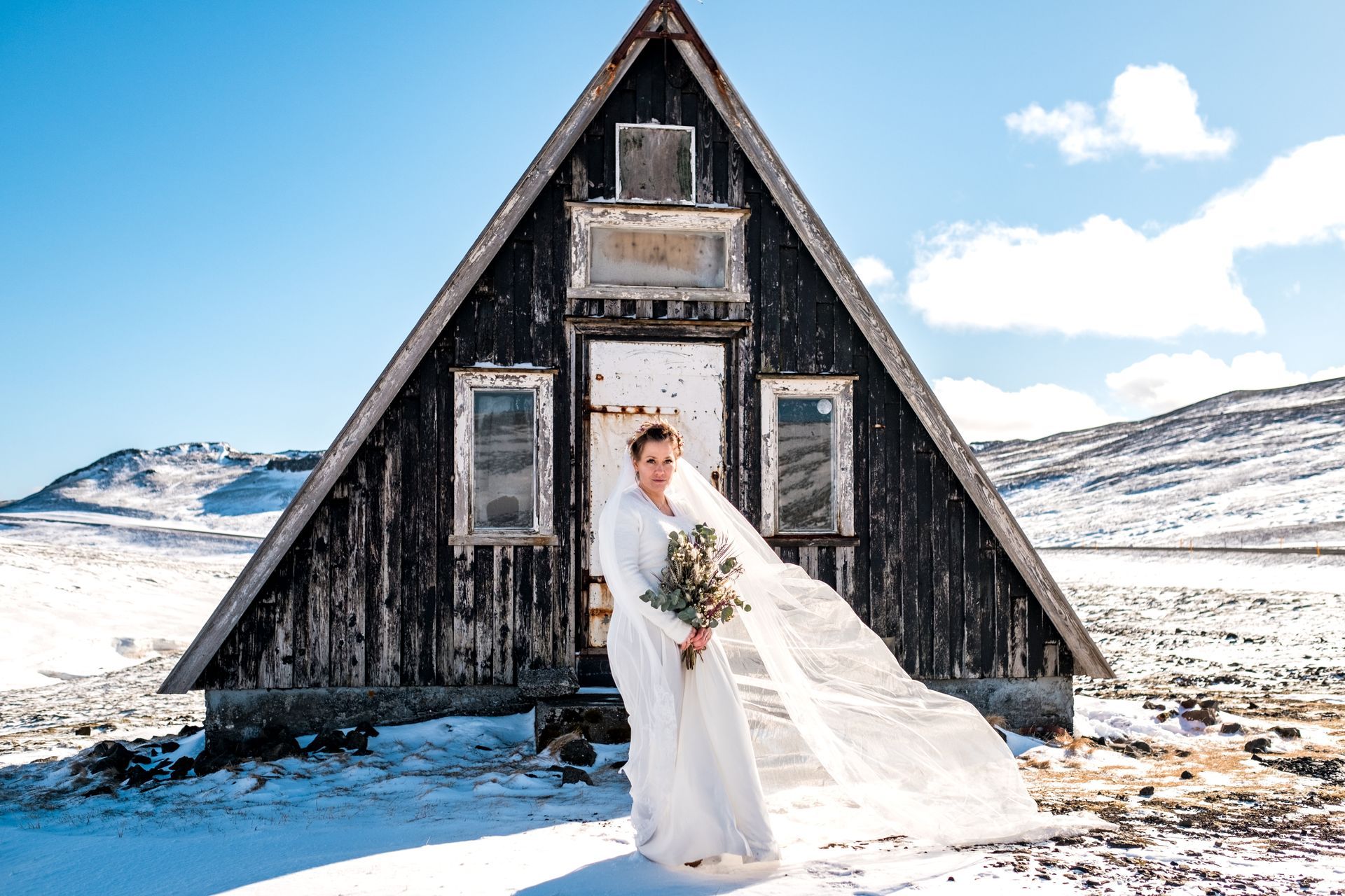 A bride in a wedding dress is standing in front of an a frame cabin in the snow.