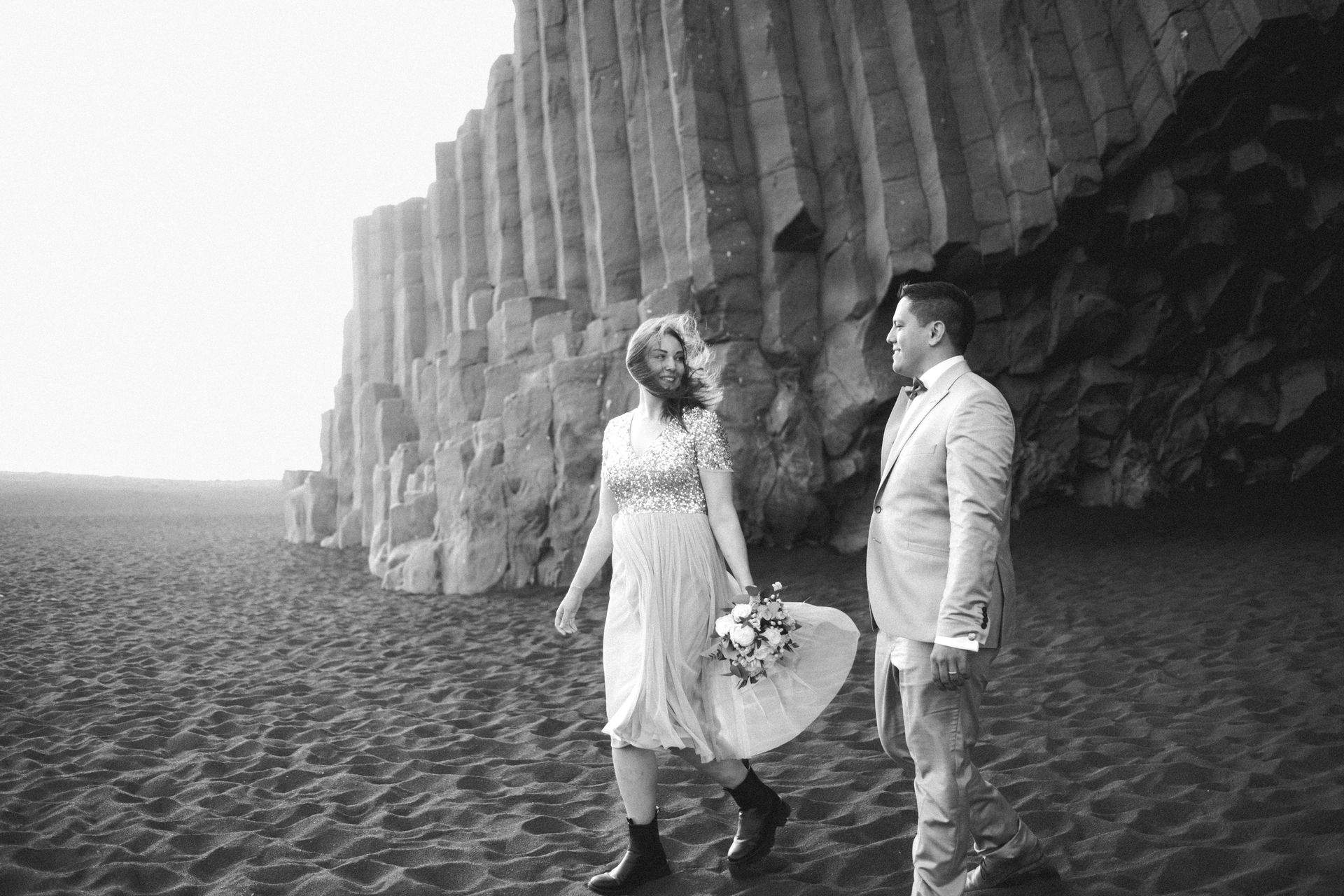 A black and white photo of a bride and groom walking on a beach.