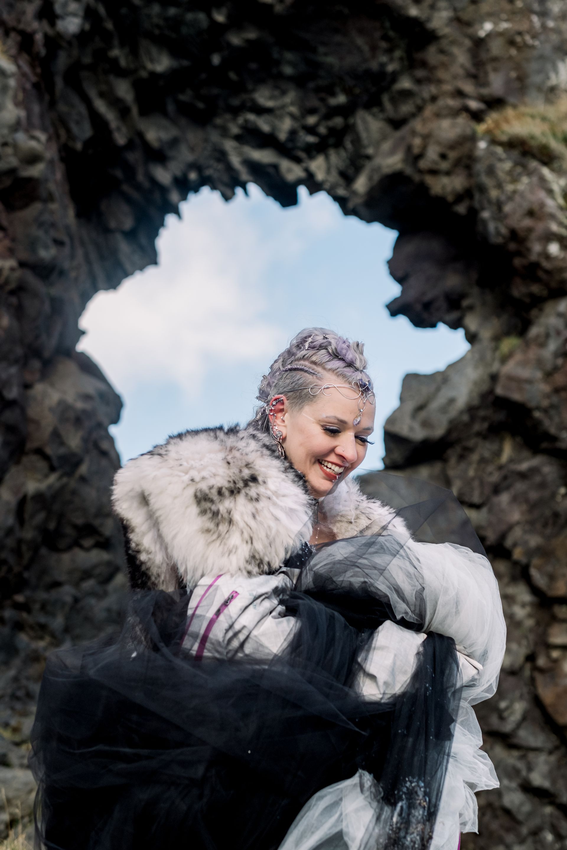 A bride in a fur-trimmed coat poses with a jeweled accessory and patterned fabric under a rock arch on a cloudy day.