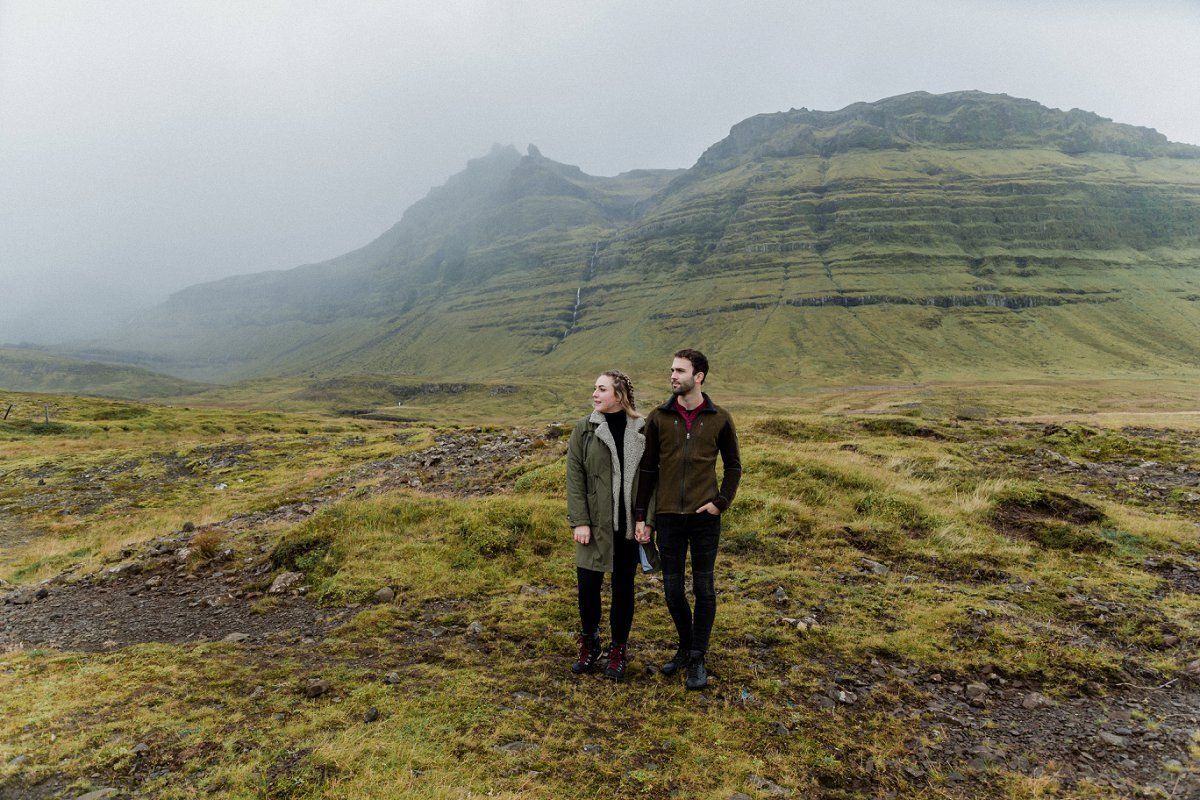A man and a woman are standing in a field with mountains in the background.