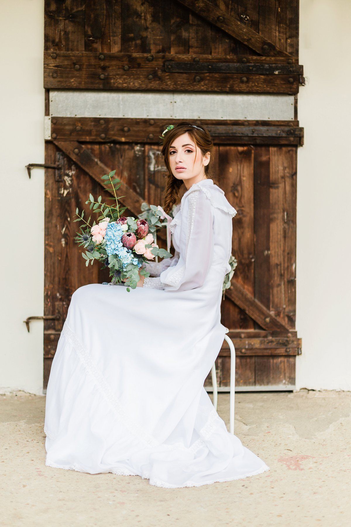 A woman in a white dress is sitting on a stool holding a bouquet of flowers.