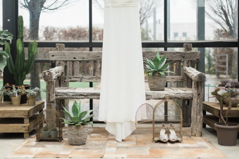 A wedding dress is hanging on a wooden bench in a greenhouse.