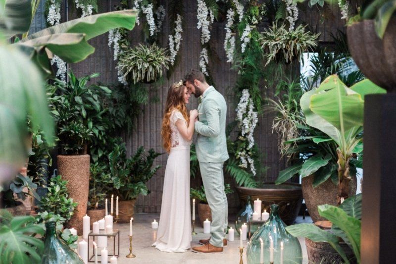 A bride and groom are kissing in a greenhouse surrounded by plants and candles.