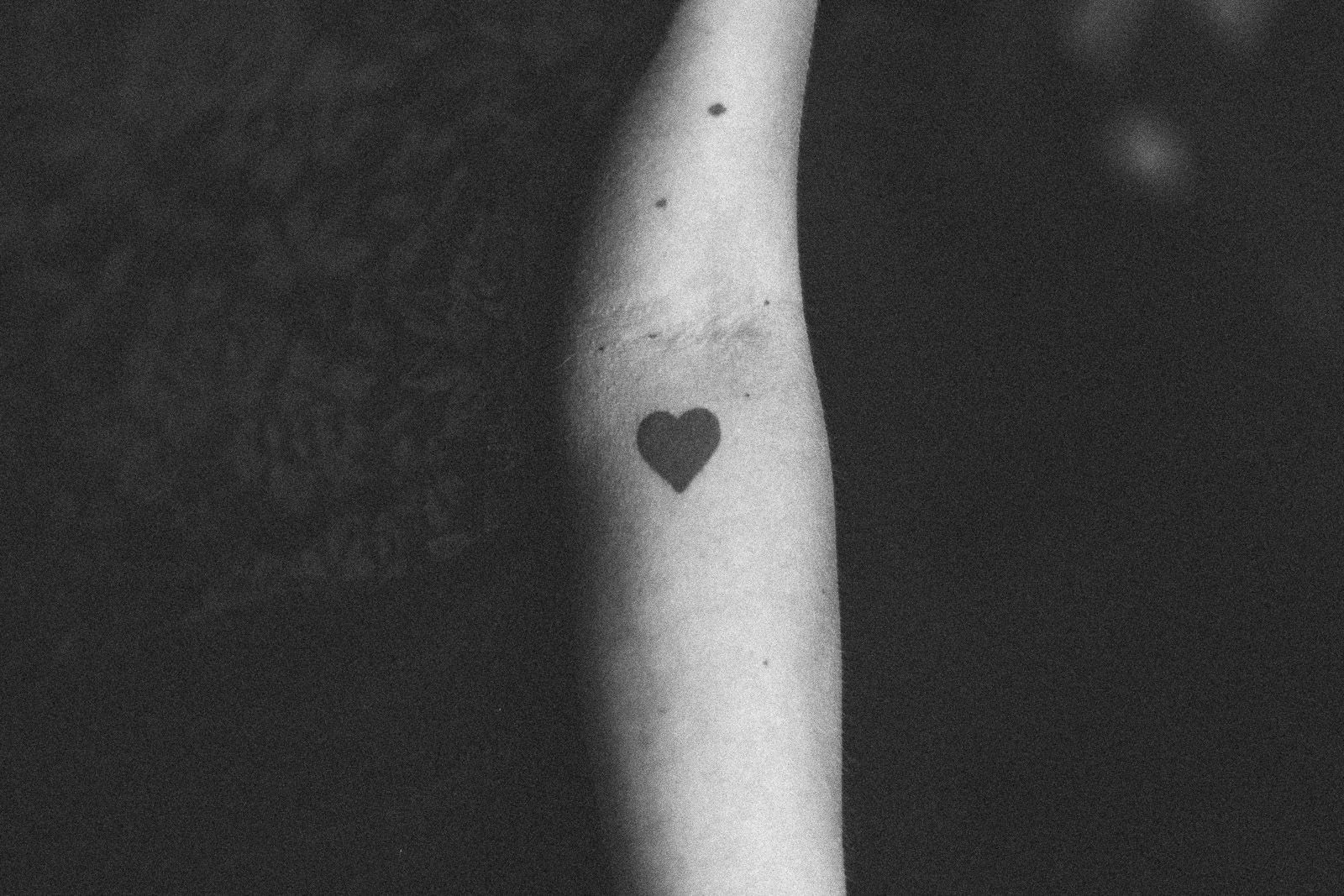 A black and white photo of a person 's arm with a small heart tattoo.