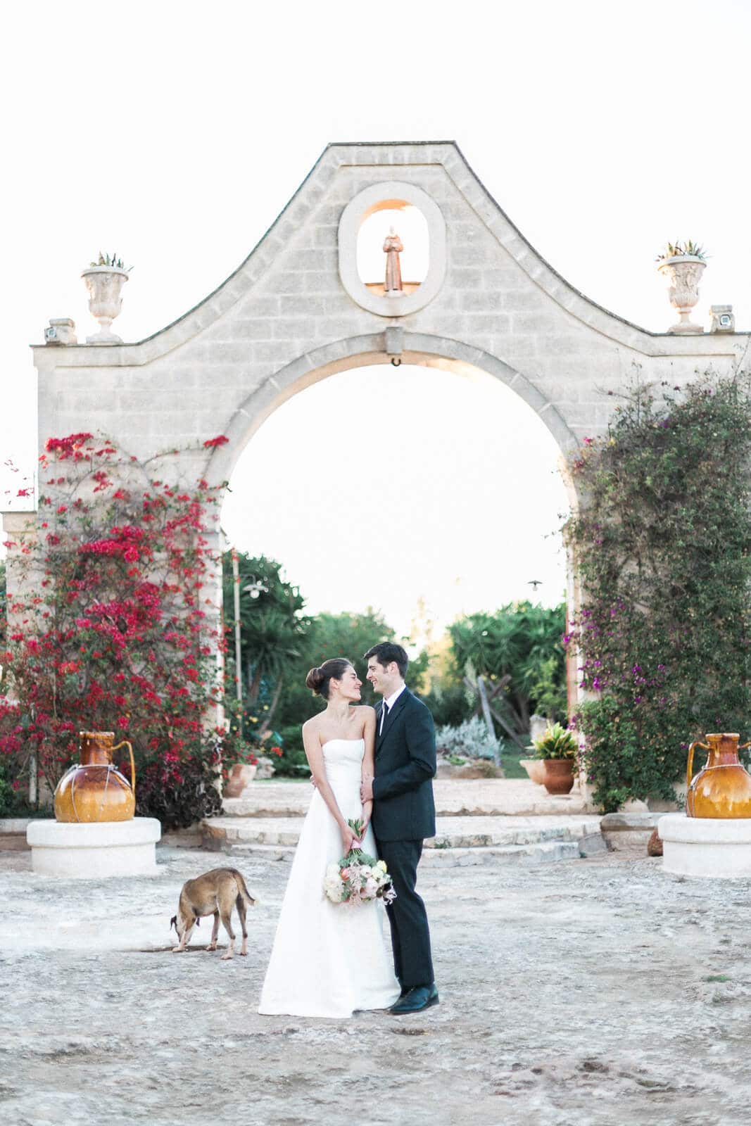 A bride and groom are posing for a picture with a dog in front of an archway.
