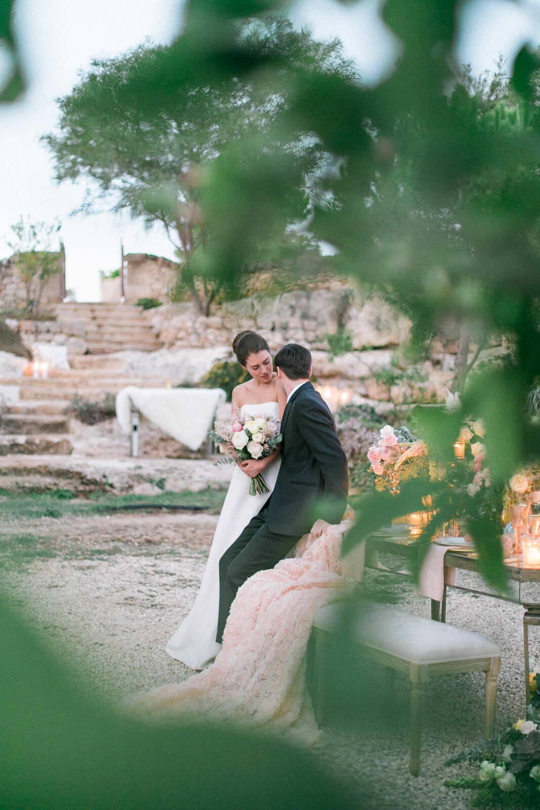 A bride and groom are sitting on a chair under a tree.