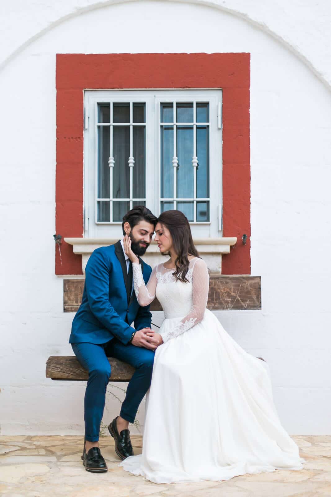 A bride and groom are sitting on a bench in front of a window.