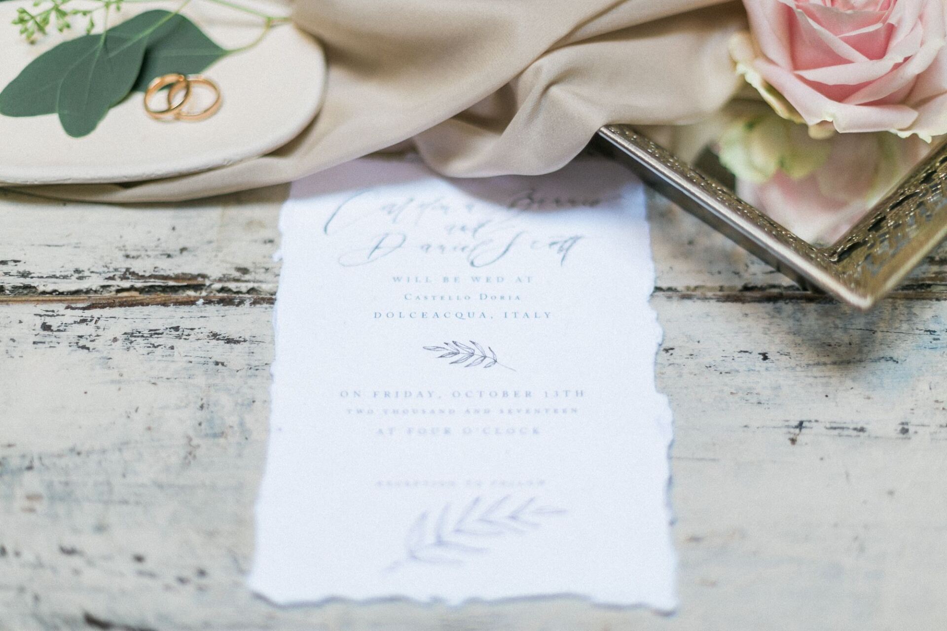 A wedding invitation is sitting on a wooden table next to a rose and wedding rings.