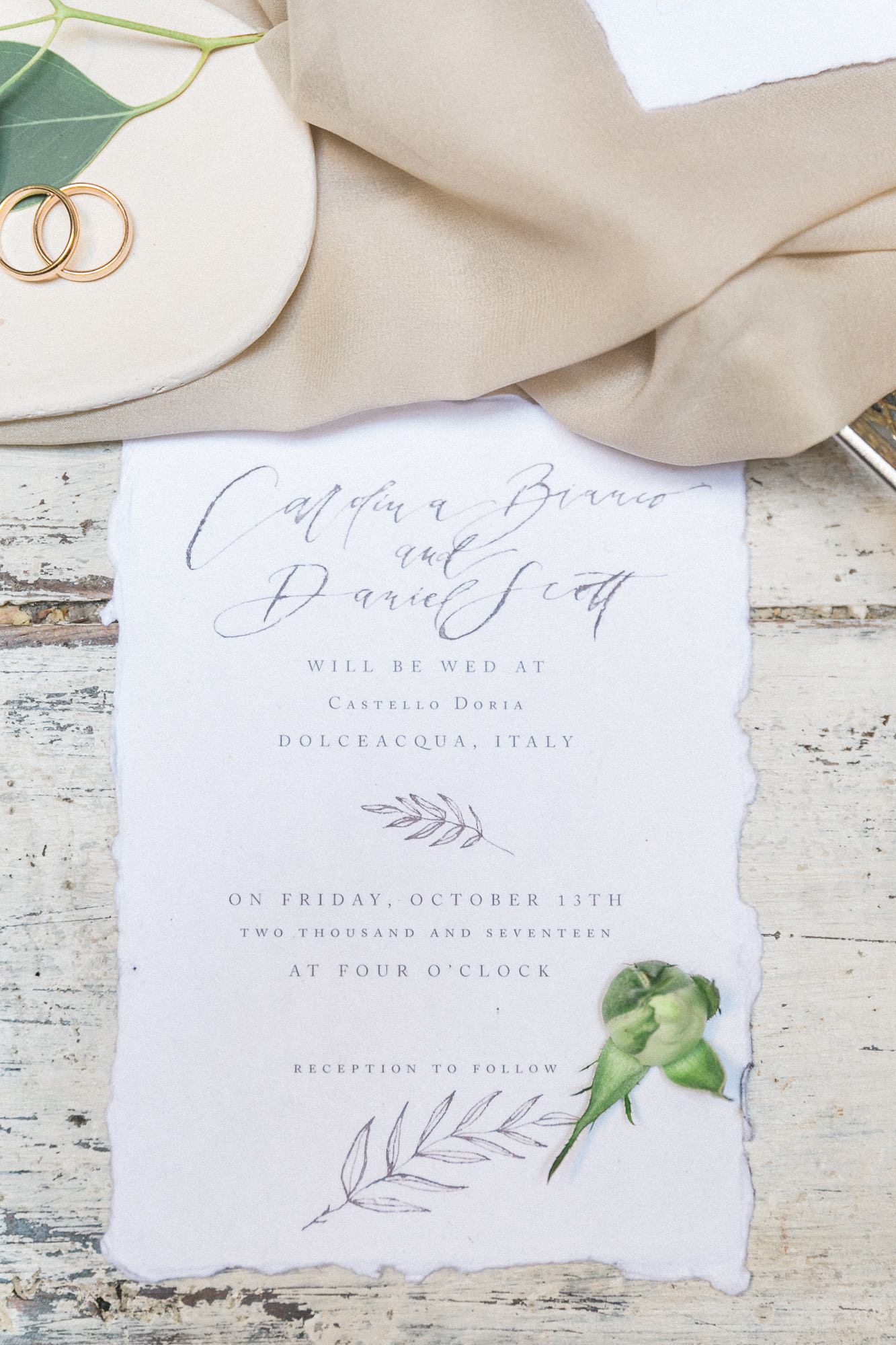 A wedding invitation is sitting on a wooden table next to a pair of wedding rings.