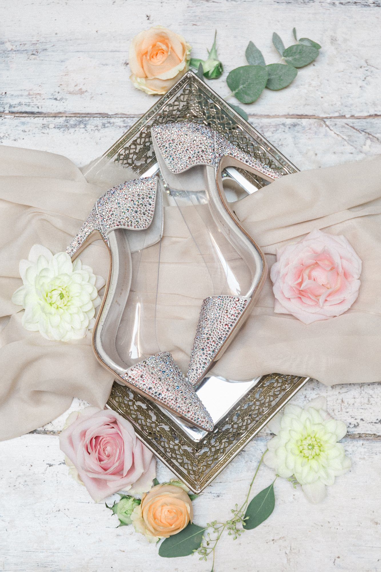 A pair of wedding shoes sitting on top of a mirror surrounded by flowers.