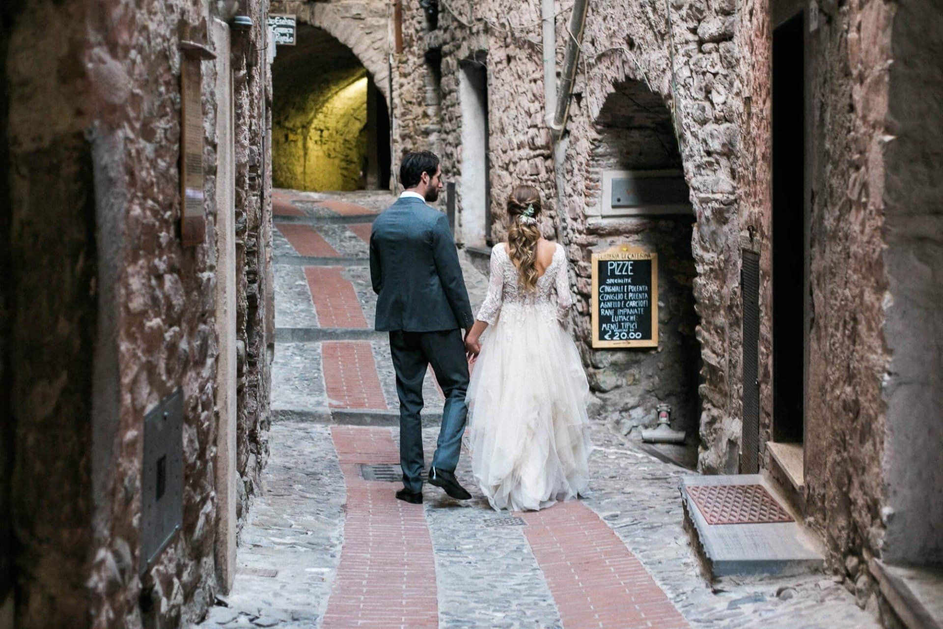 A bride and groom are walking down a narrow street holding hands.