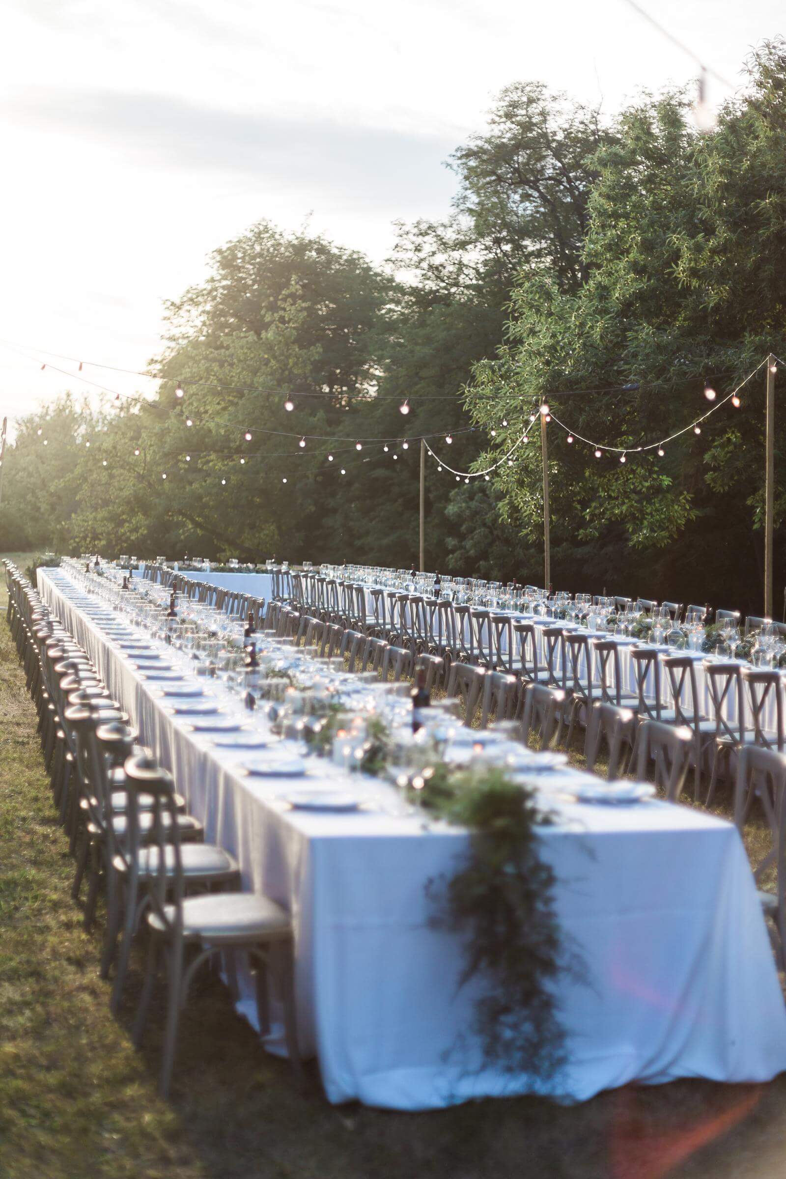 A very long table with white linens and chairs in a field.