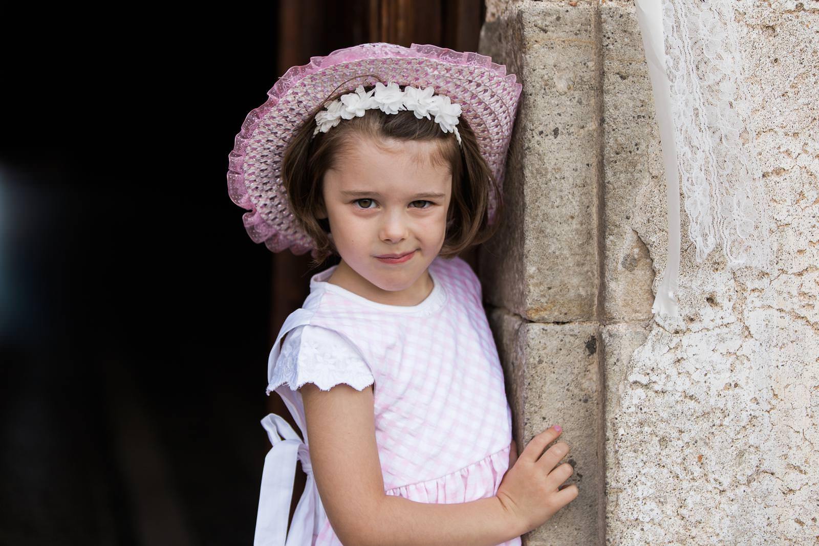 A little girl wearing a pink hat and a white dress is leaning against a stone wall.
