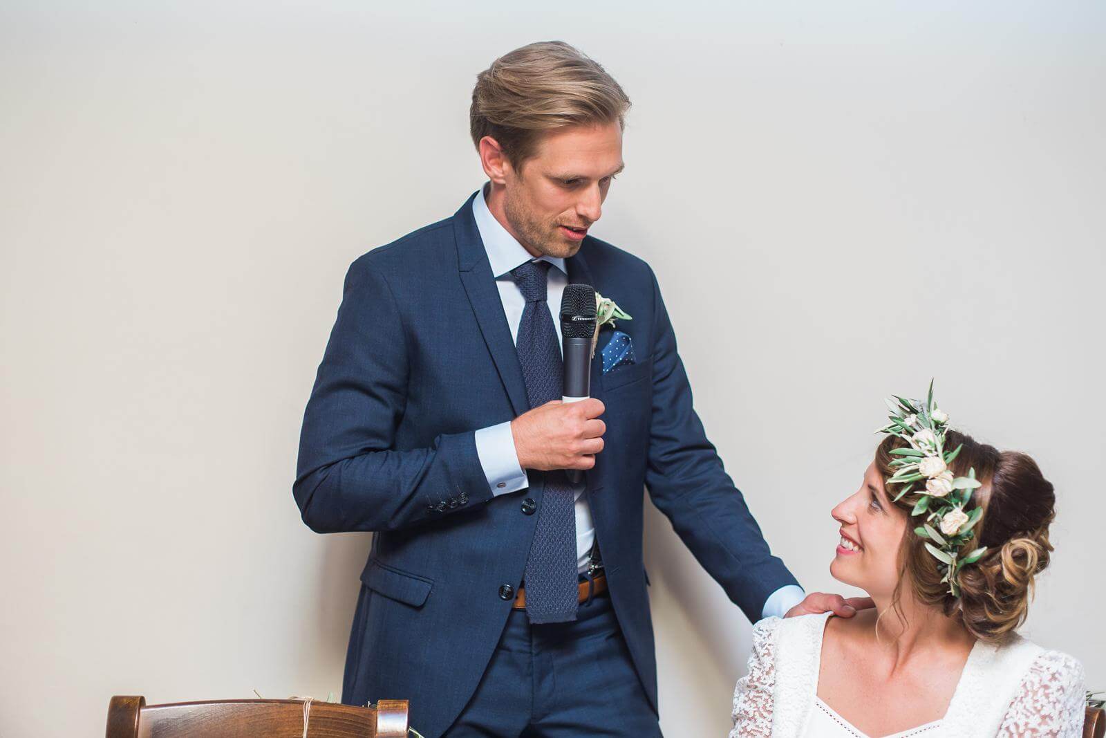 A bride and groom are sitting at a table while the groom is giving a speech.