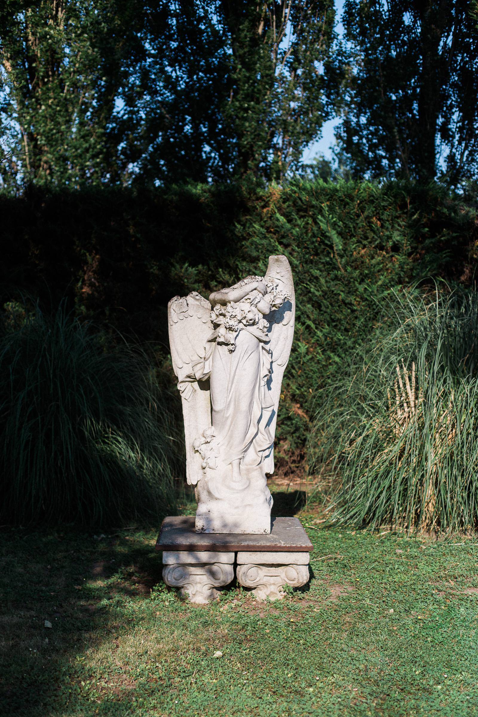 A statue of an angel is sitting in the grass in front of a hedge.