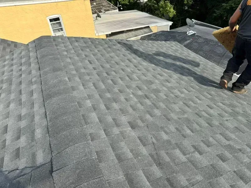 Dark gray shingled roof with a worker standing at the edge near a yellow building
