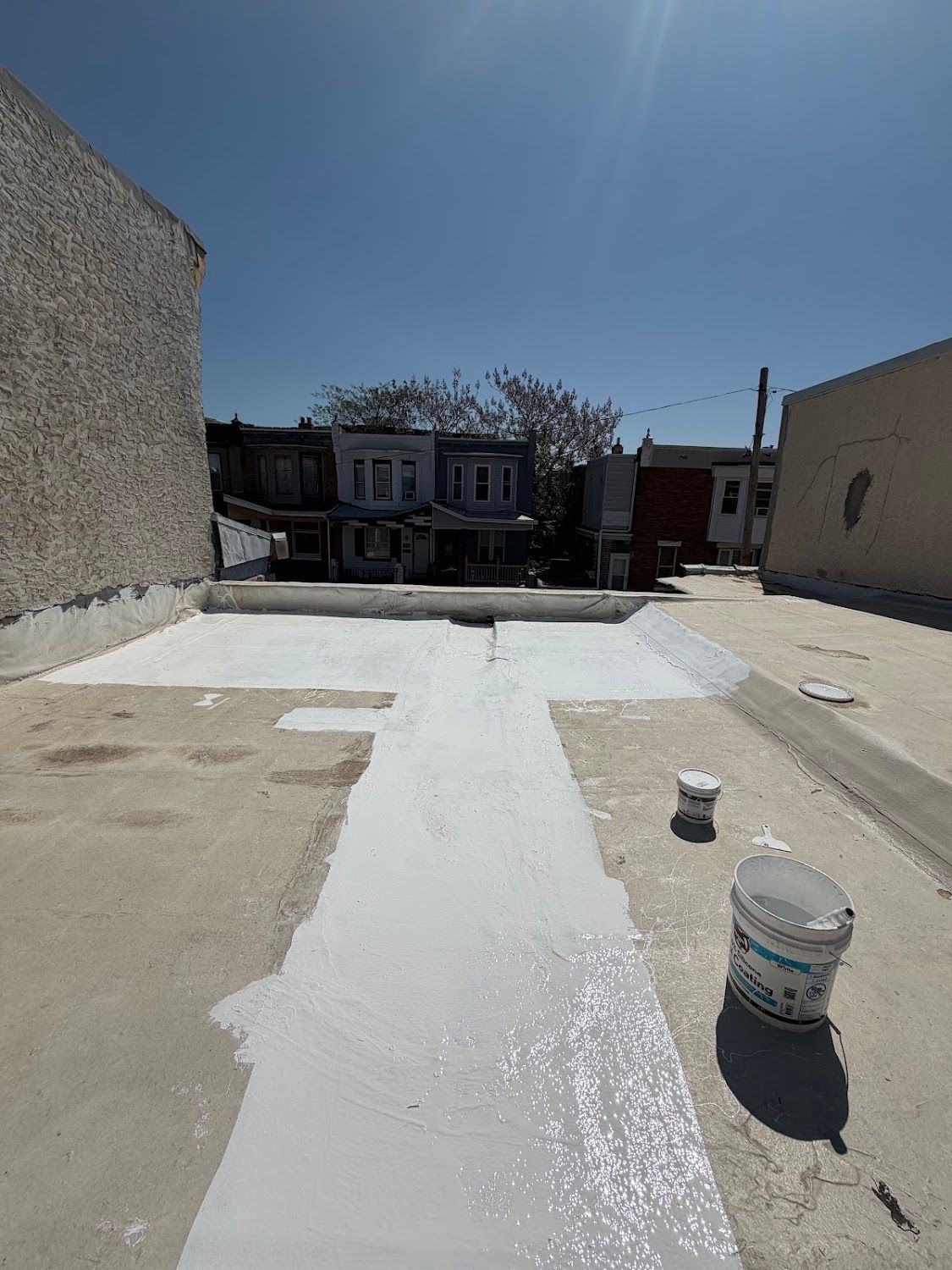 Rooftop with freshly coated white roof surface, buckets of paint, and neighboring houses under a clear blue sky