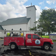 Red utility truck parked outside a white church with a tall steeple on a cloudy day.