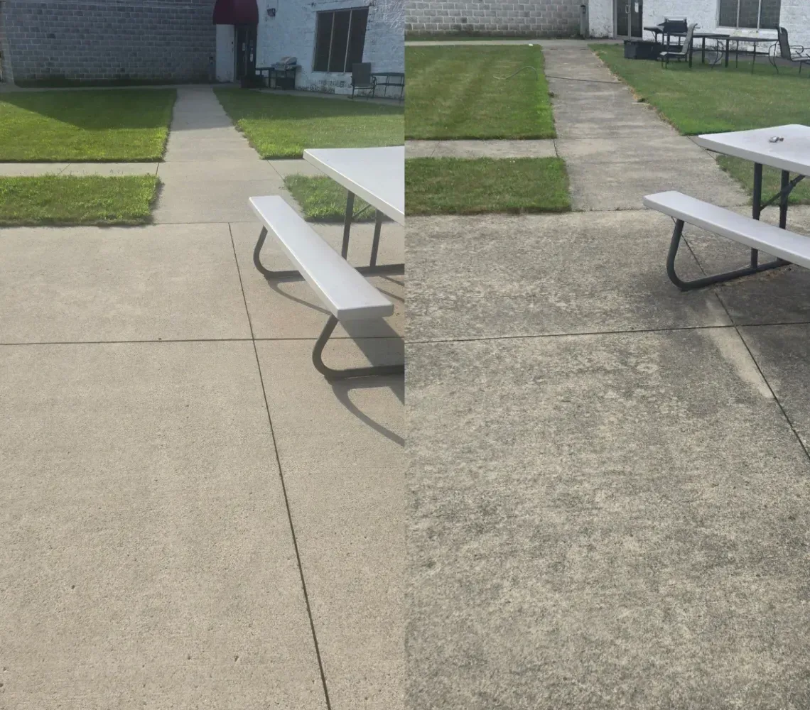 Concrete patio with picnic tables and white benches beside a grassy courtyard and brick building.