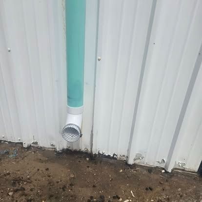 White corrugated wall with a turquoise downspout ending near the ground beside dirt and gravel.
